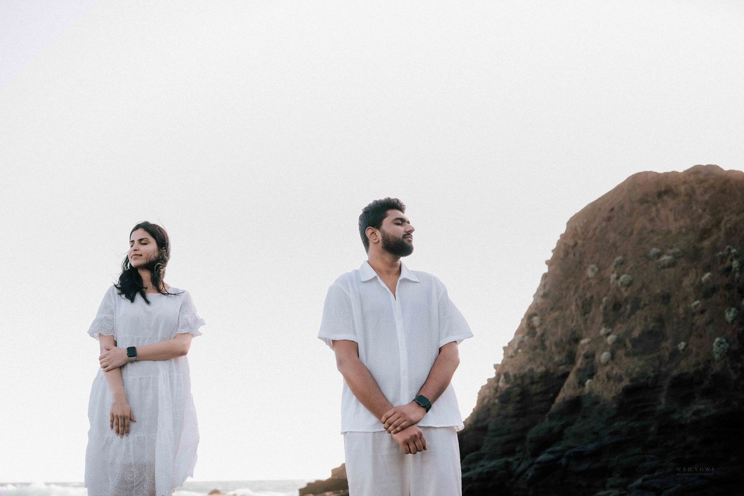 Couple in white outfits standing near a large rock looking upward by the ocean.