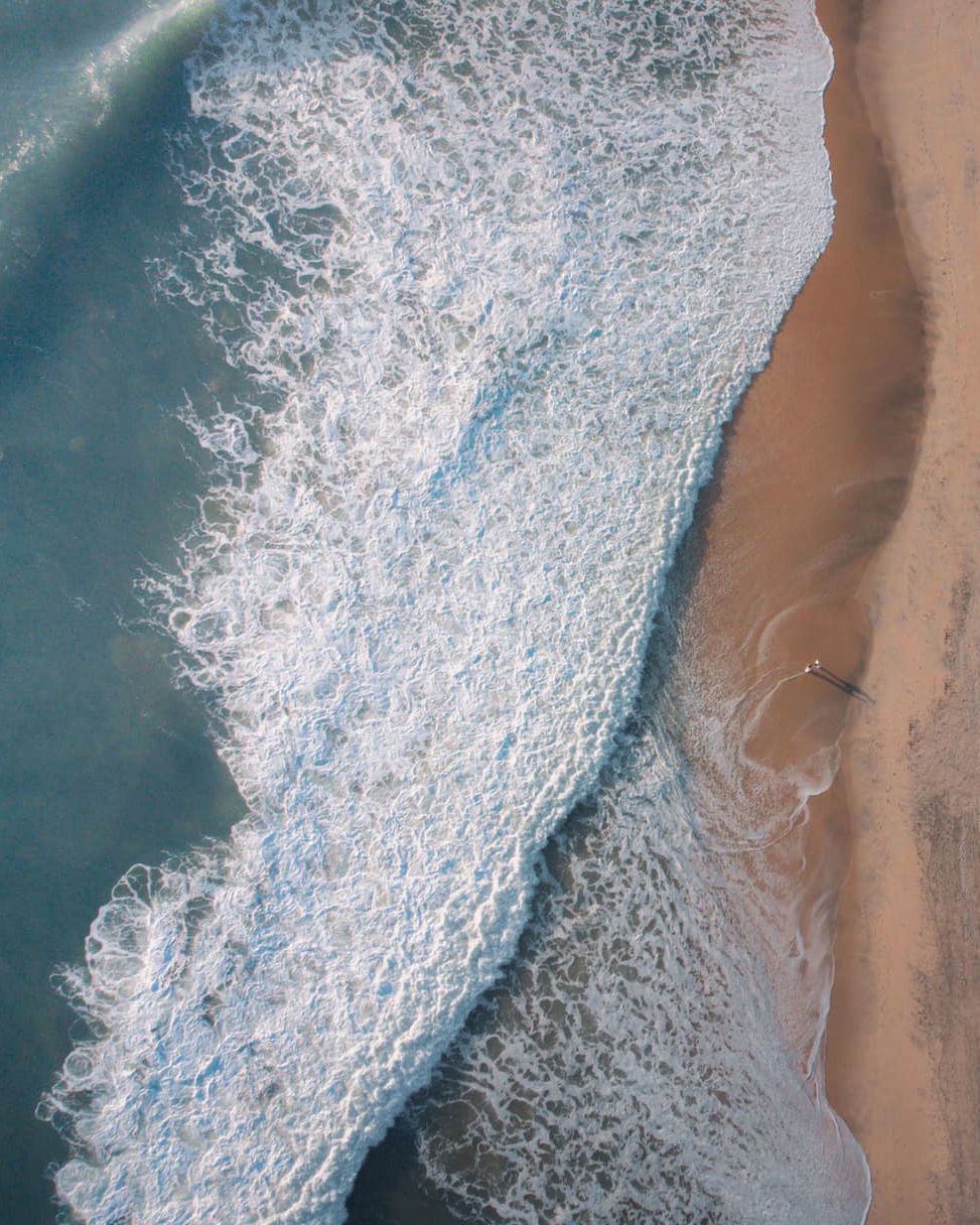 Aerial drone view of foamy ocean waves crashing onto sandy beach shoreline with turquoise water.