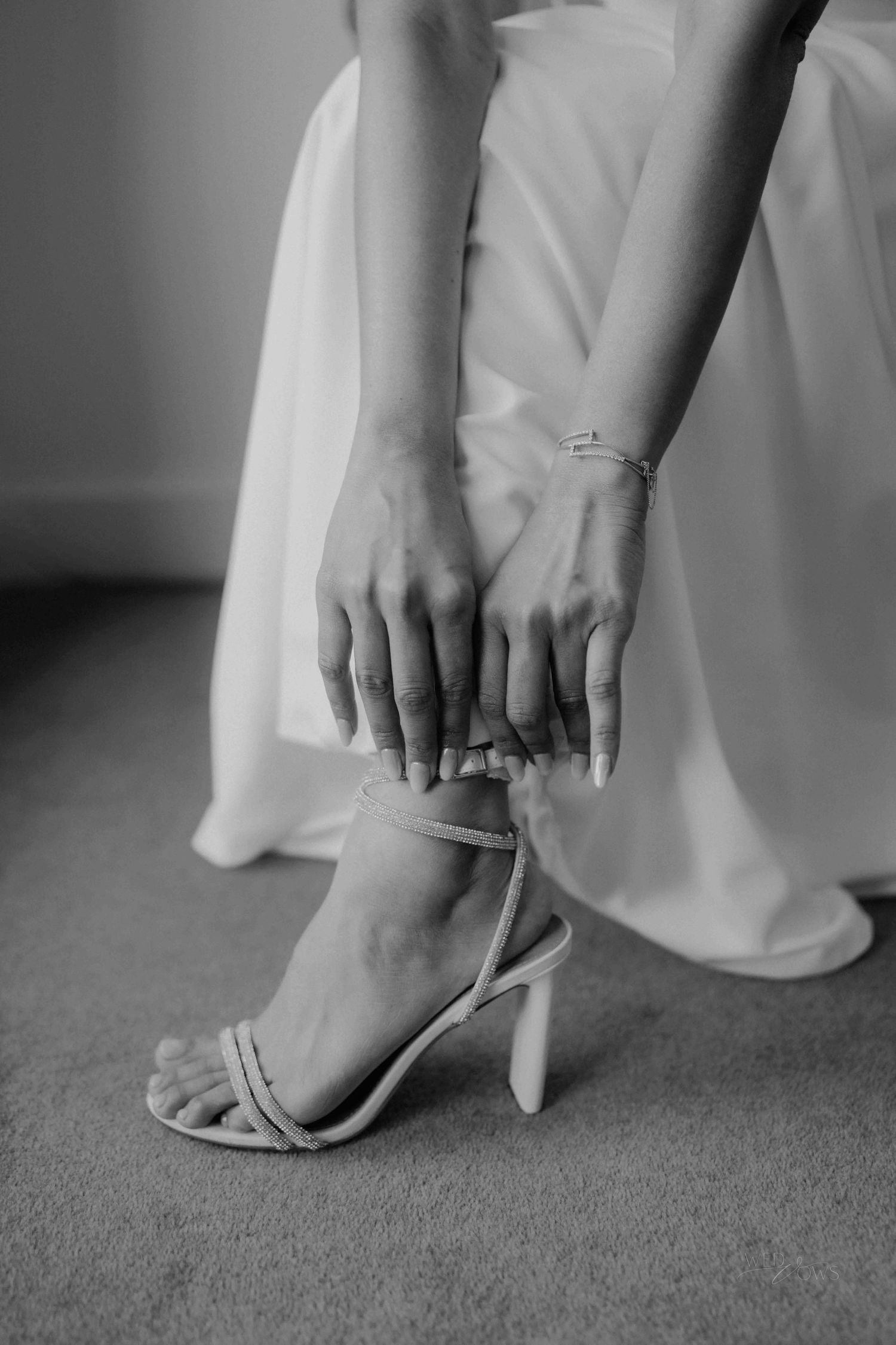 Black and white close-up of bride fastening strappy stiletto heel, showing bracelet and manicure.