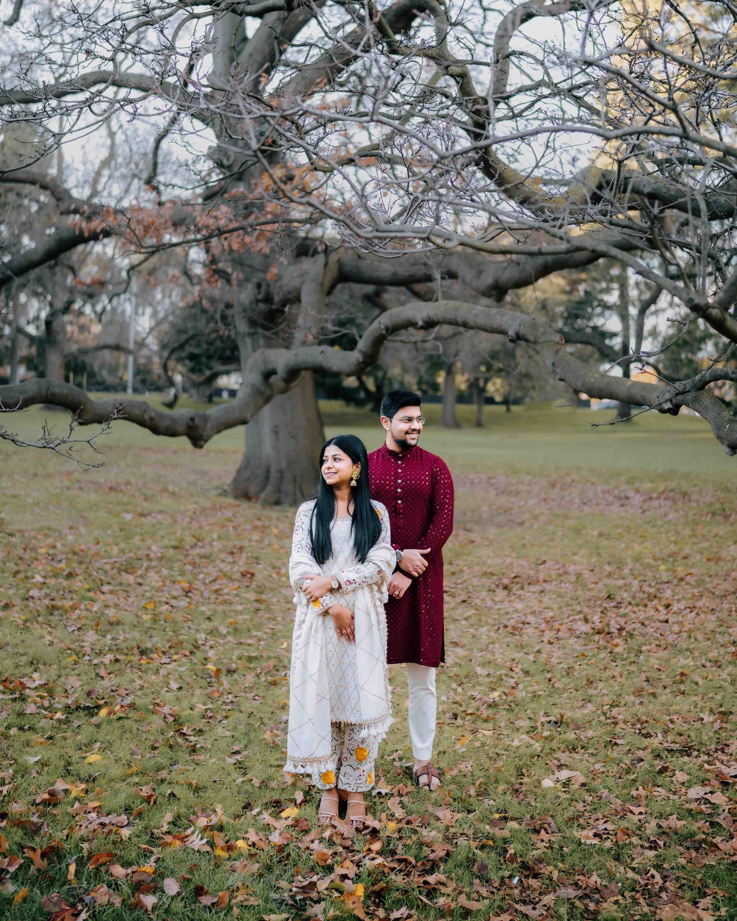 Couple in traditional South Asian attire standing in autumn park with bare trees and fallen leaves.