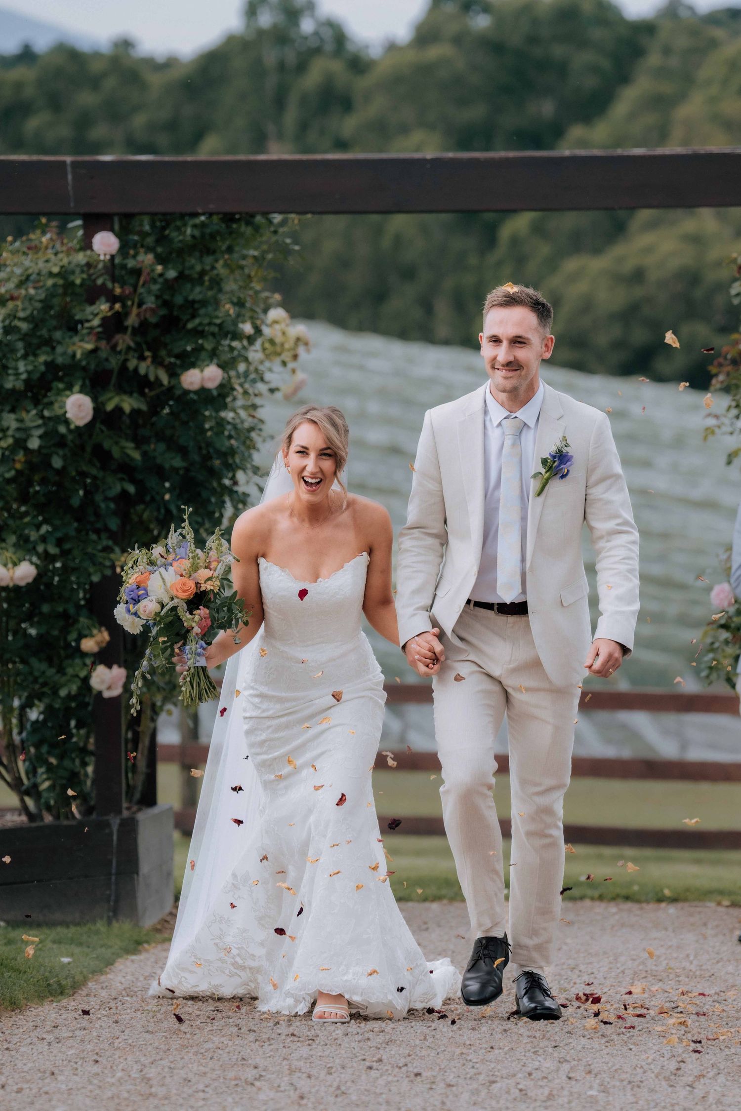 Joyful newlywed couple walking down aisle with confetti, bride in white gown holding colorful bouquet, groom in beige suit.