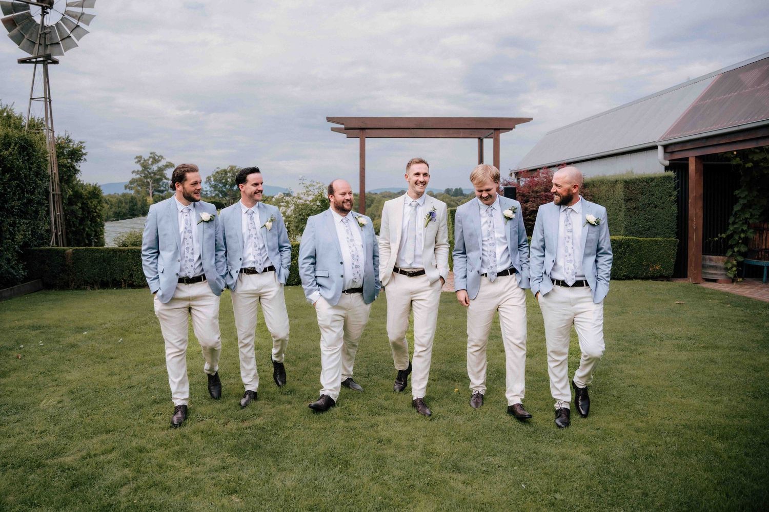 Groomsmen in light blue suits and cream pants laughing while walking together on green grass.