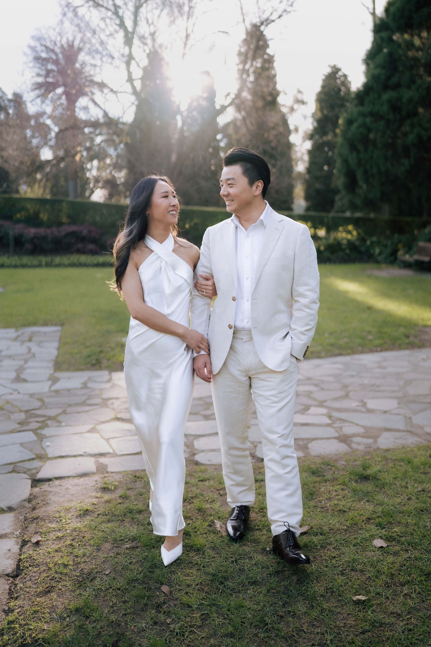 Couple in matching white outfits walking hand in hand through a sunlit garden, smiling at each other.