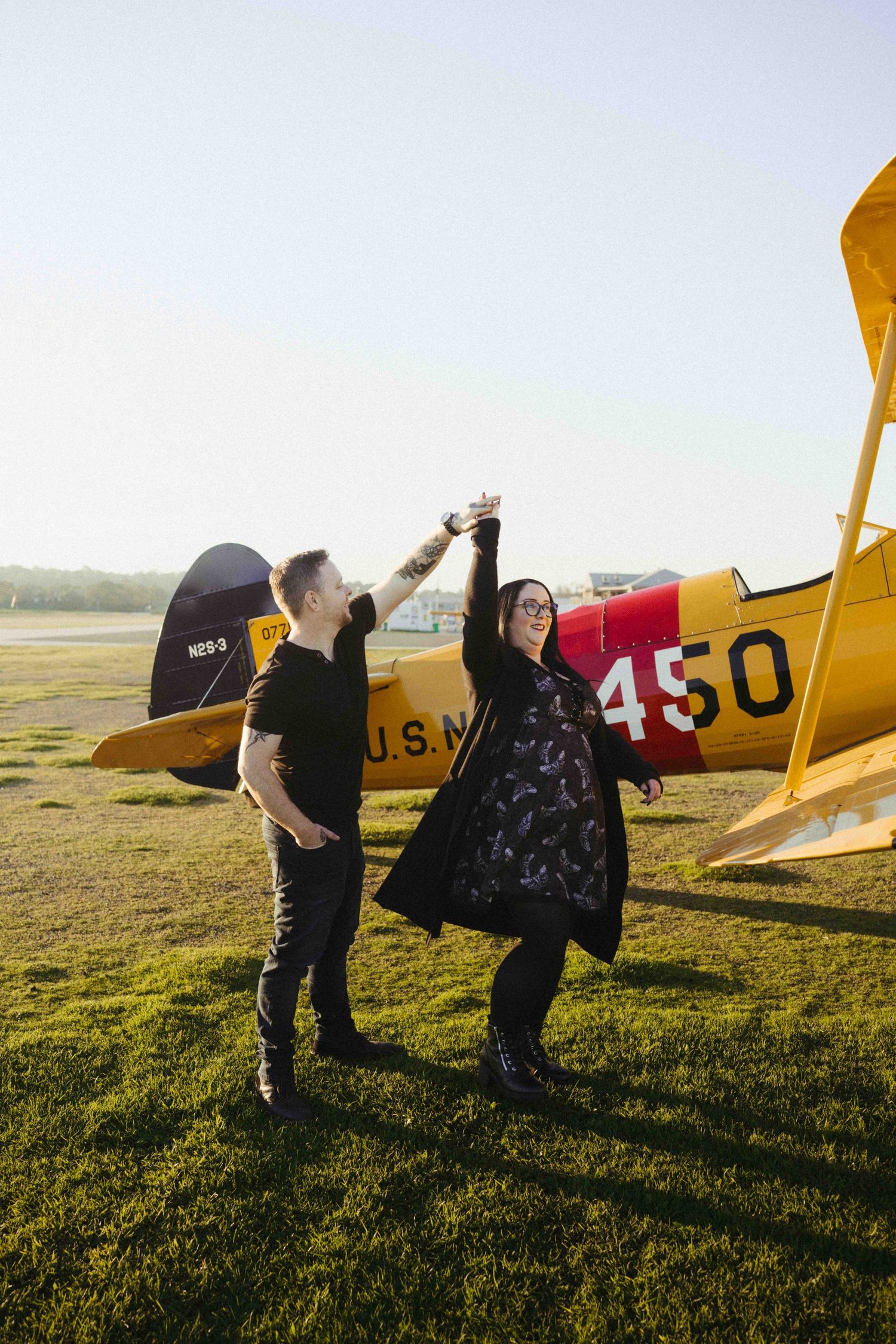 Couple dancing joyfully in front of a vintage yellow U.S. Navy biplane numbered 450 on a sunny airfield.