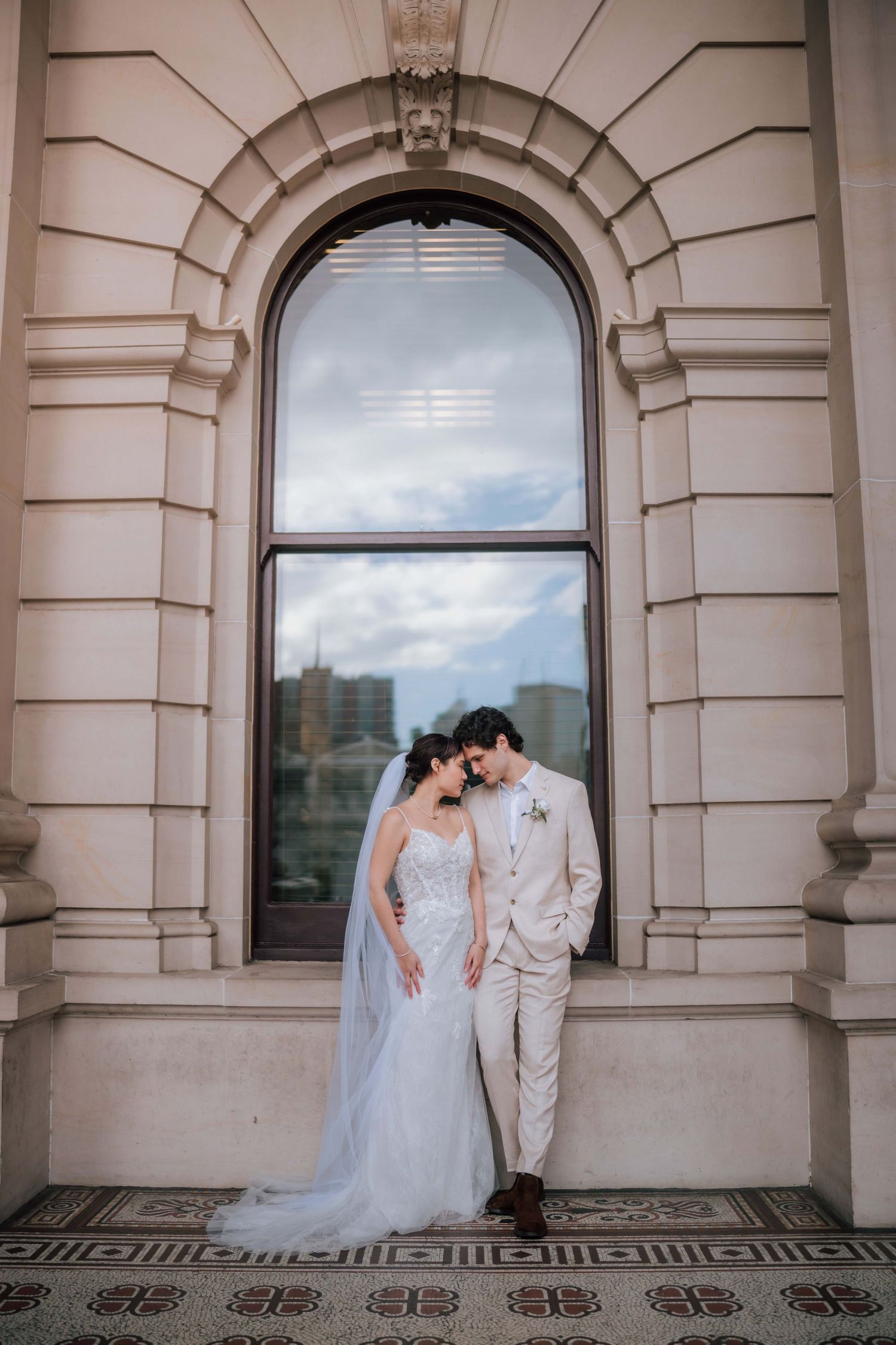 Bride and groom sharing intimate moment beneath arched window on ornate building exterior.