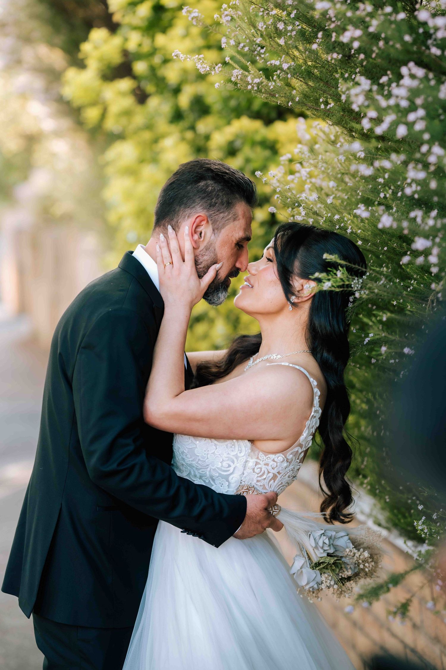 Bride and groom sharing a romantic moment about to kiss near blooming flowers outdoors.