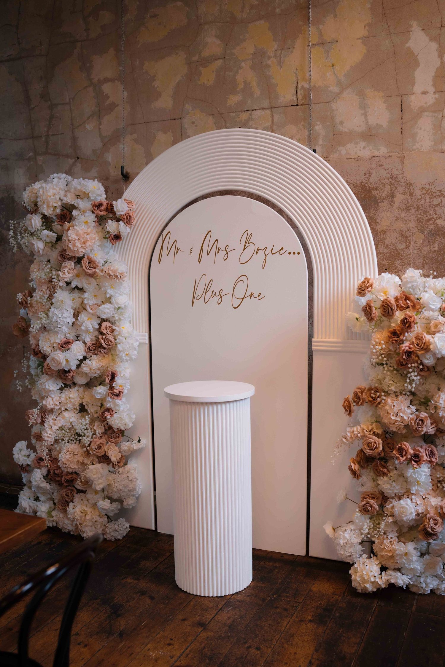 White ribbed arch wedding sign reading Mr and Mrs Bosie Plus One flanked by lush blush and white floral arrangements.