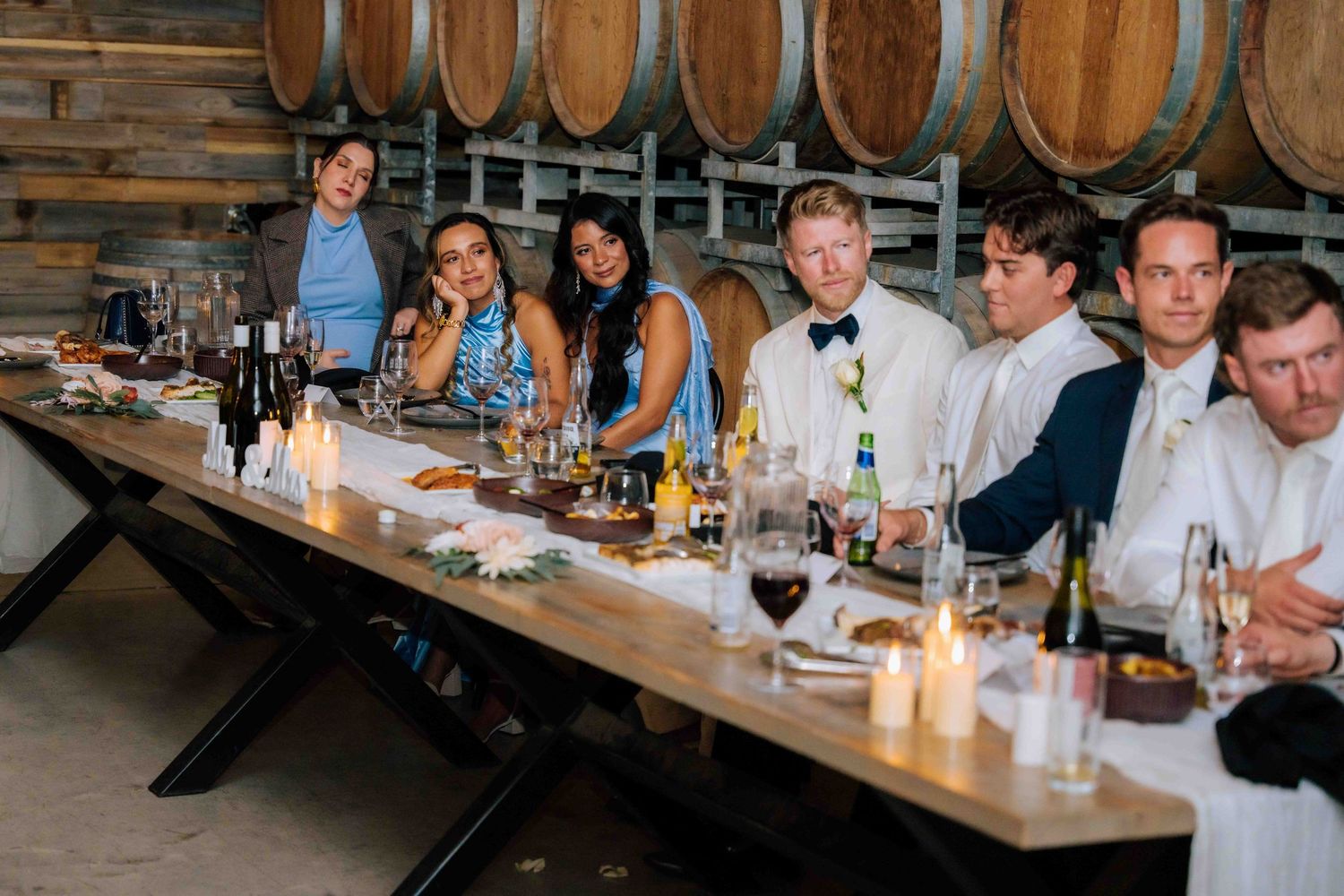 Wedding guests seated at long reception table in rustic winery barrel room with candles.
