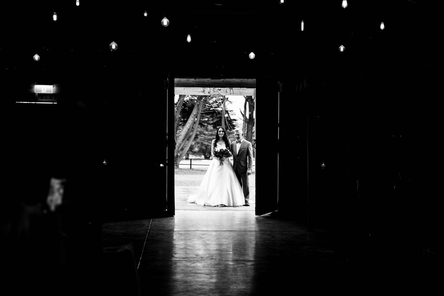Bride and groom silhouetted in doorway of dark wedding venue in black and white photo.