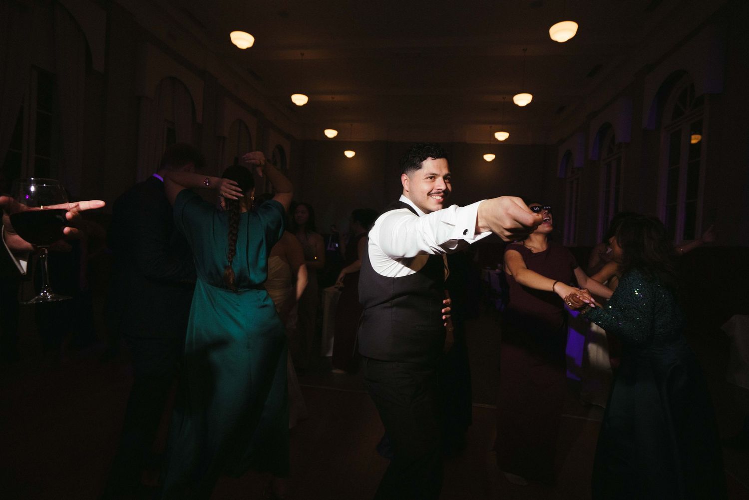 Groom in white shirt dances energetically with guests on dimly lit wedding reception dance floor.