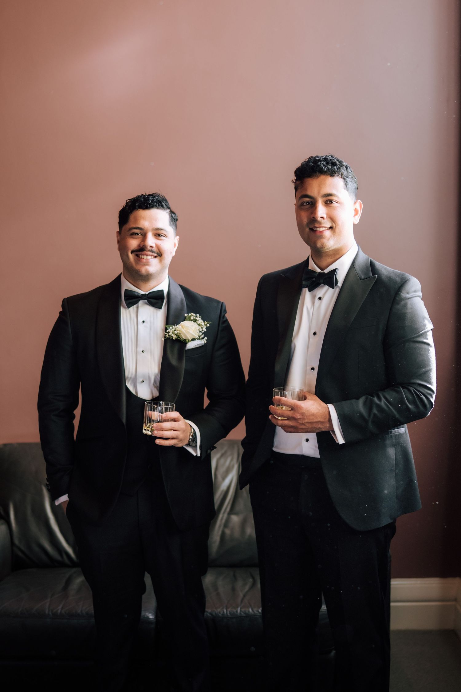 Two grooms in black tuxedos with bow ties holding drinks and smiling at their wedding celebration.