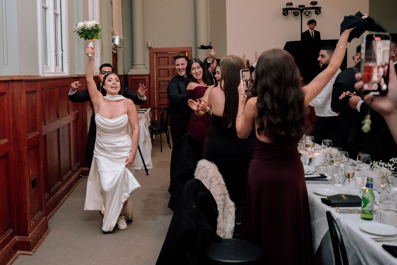 Joyful bride entering wedding reception raising bouquet as guests cheer and celebrate.