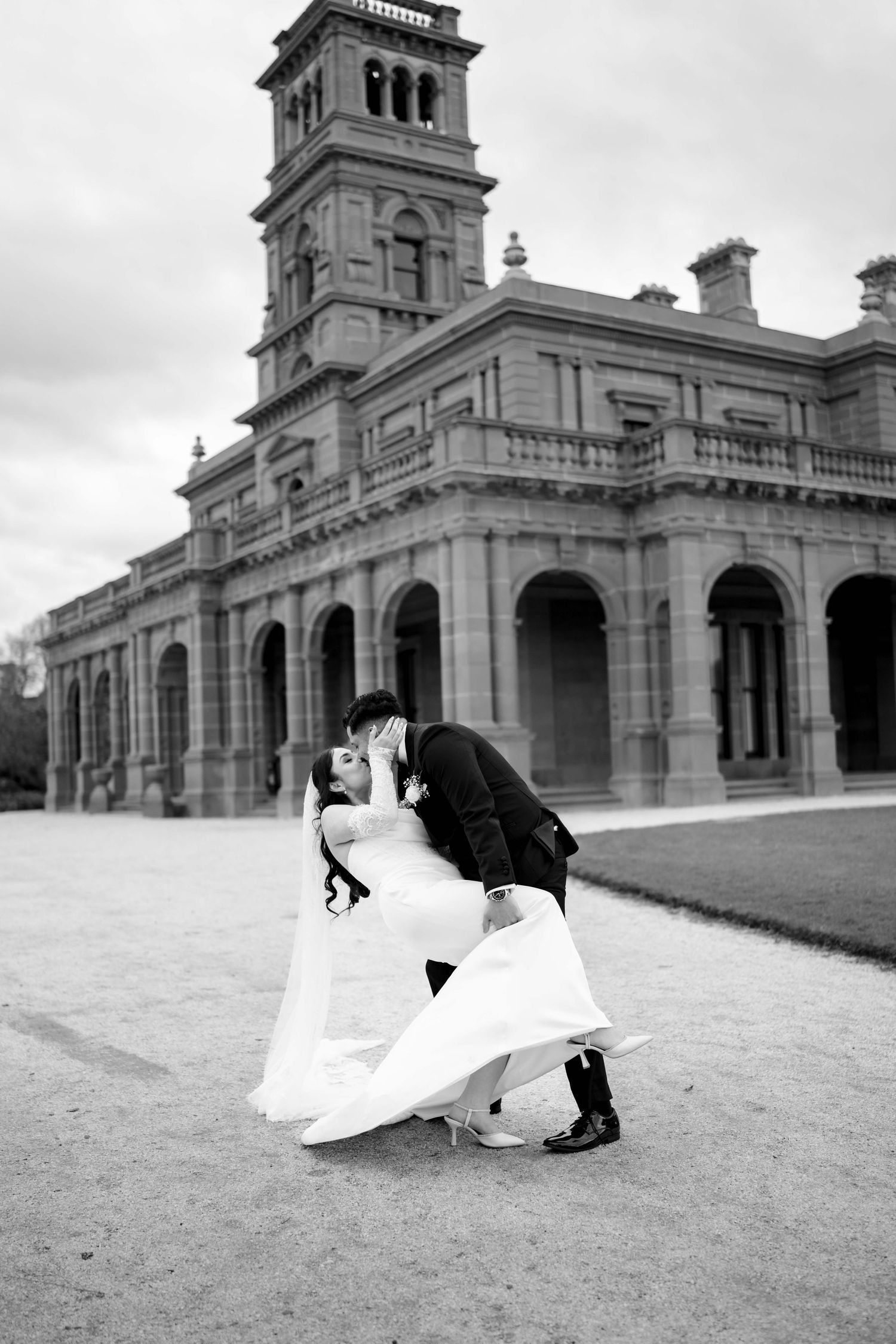 Black and white photo of bride and groom sharing a dip kiss in front of historic mansion.