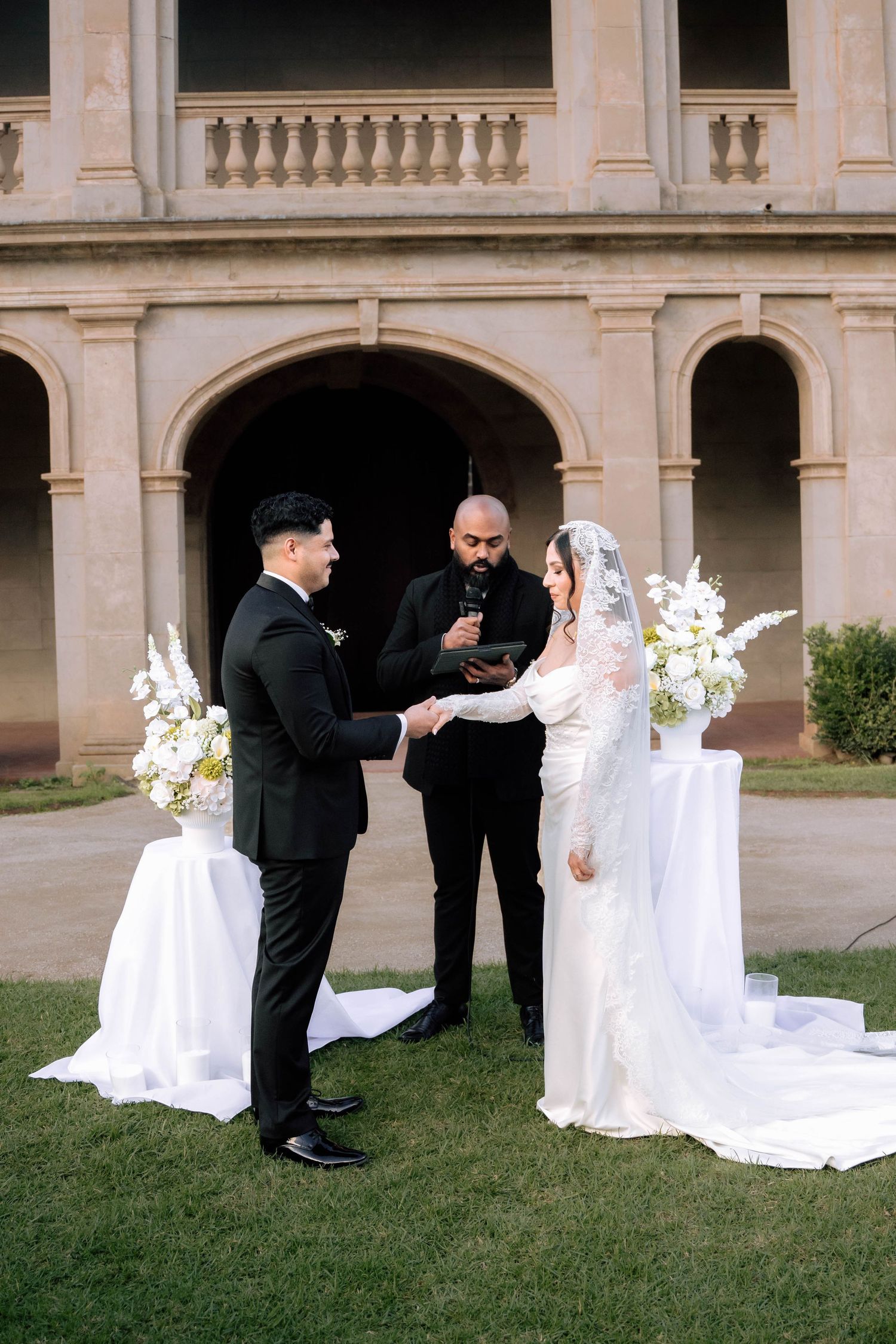 Couple exchanging vows outdoors before historic arched building with white floral arrangements.
