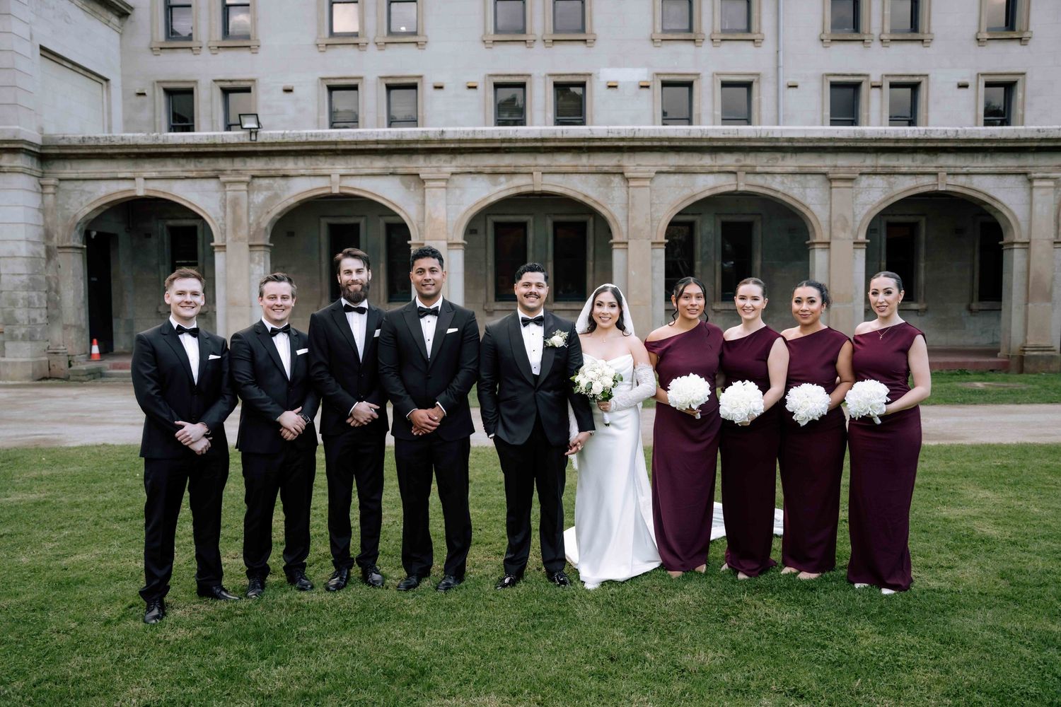 Wedding party poses in front of a historic building with groomsmen and burgundy-gowned bridesmaids.