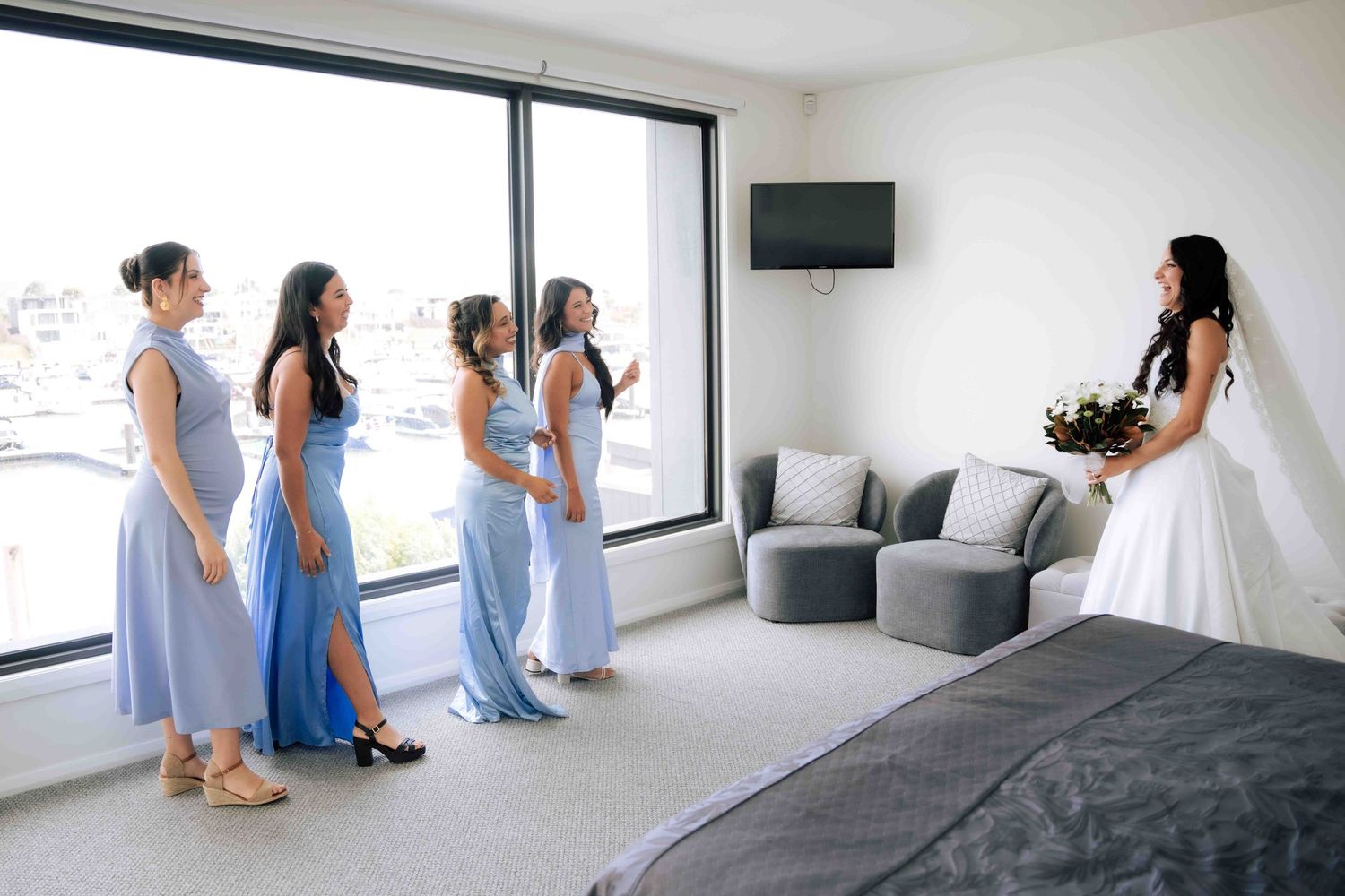 Bride in white gown surprises her four bridesmaids in blue dresses during wedding preparations in a hotel room.