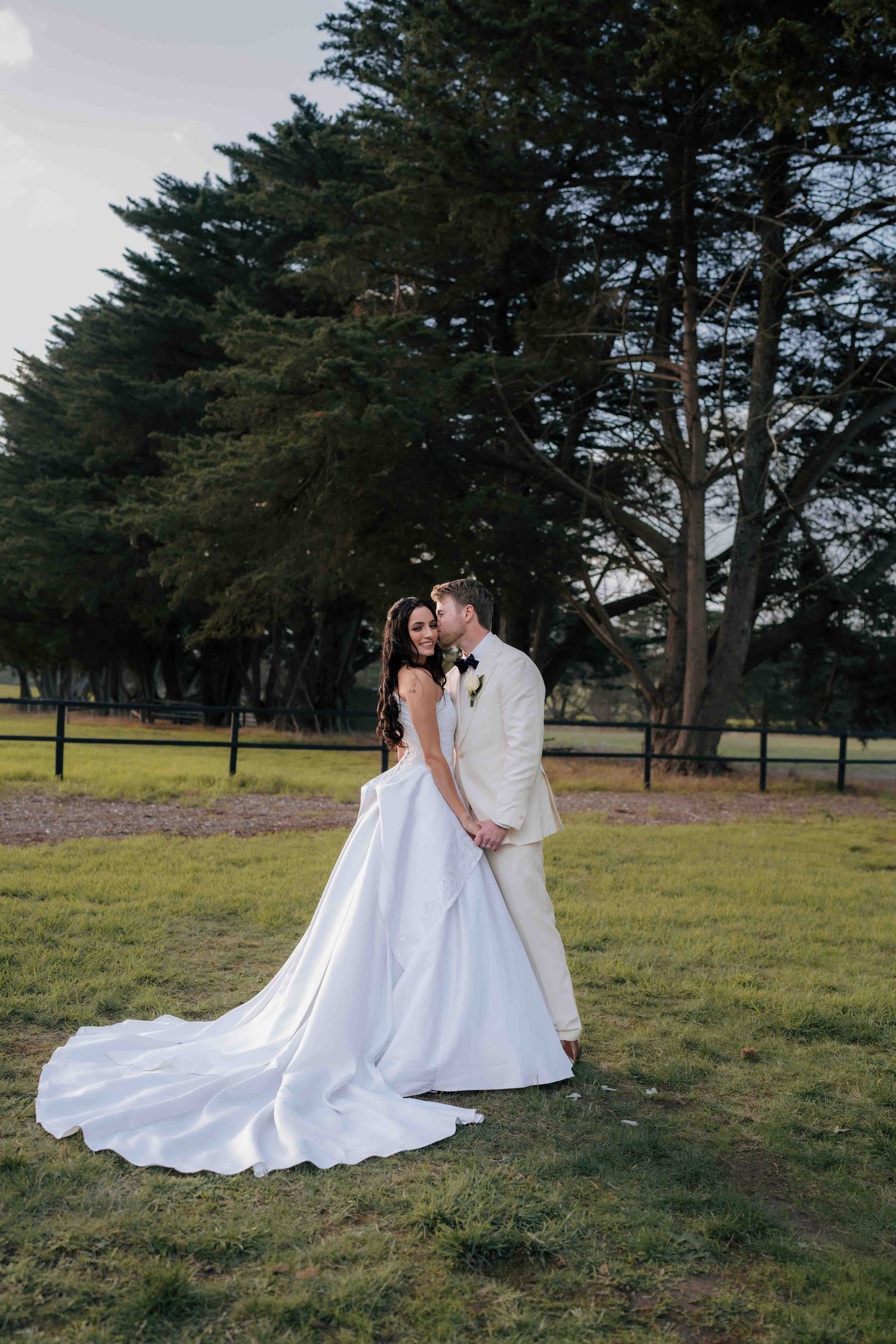 Bride in elegant white ballgown with long train poses with groom in cream suit before tall cypress trees.