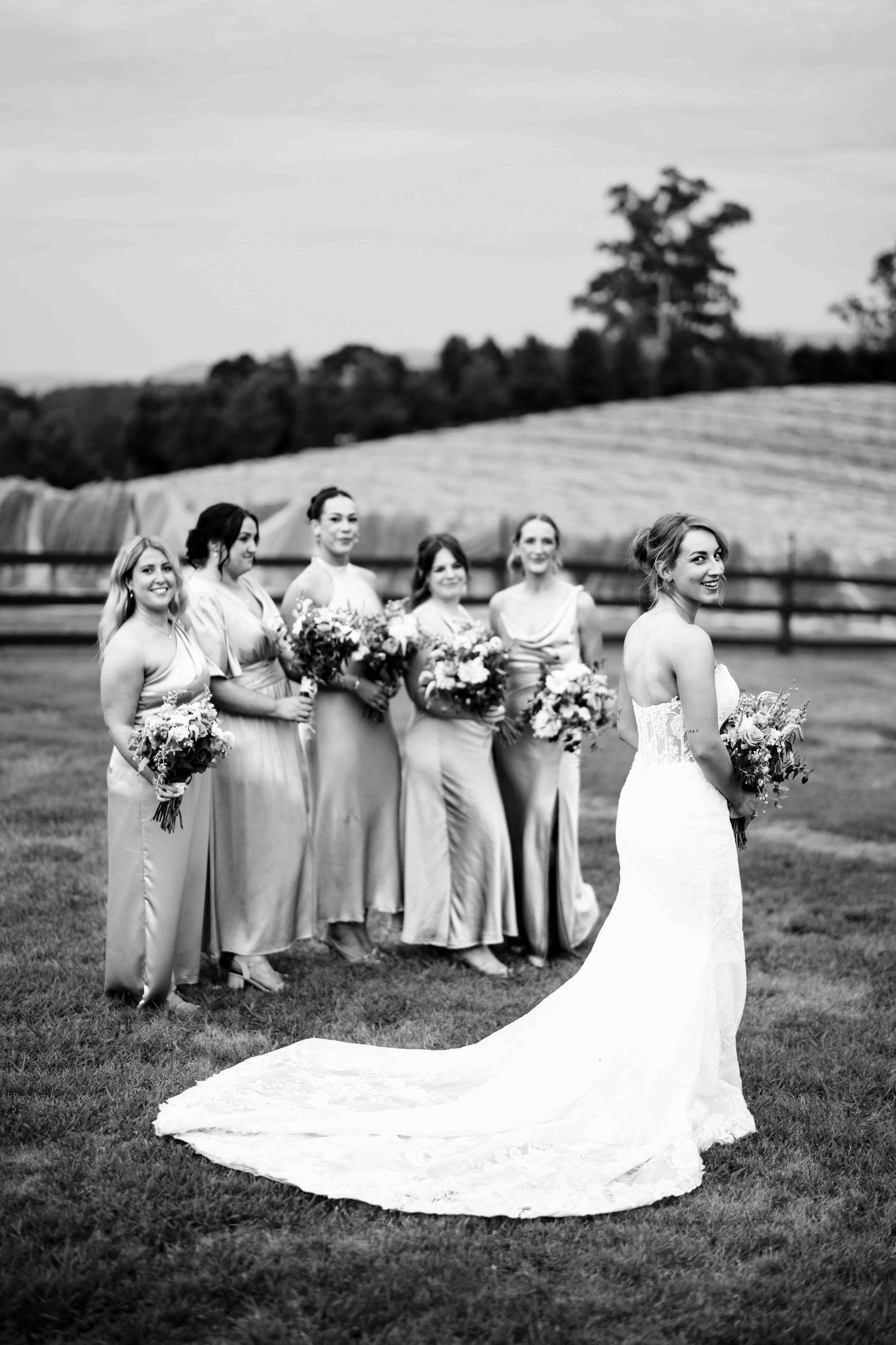 Black and white photo of bride in elegant gown with long train and five bridesmaids in satin dresses outdoors.