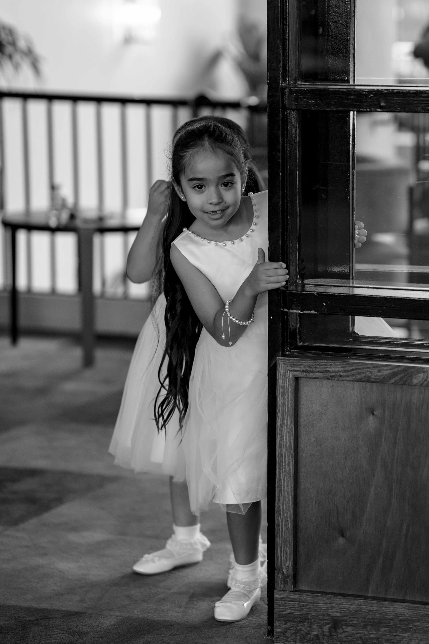 An adorable young flower girl in a white dress peeking around a wooden door at a wedding venue.