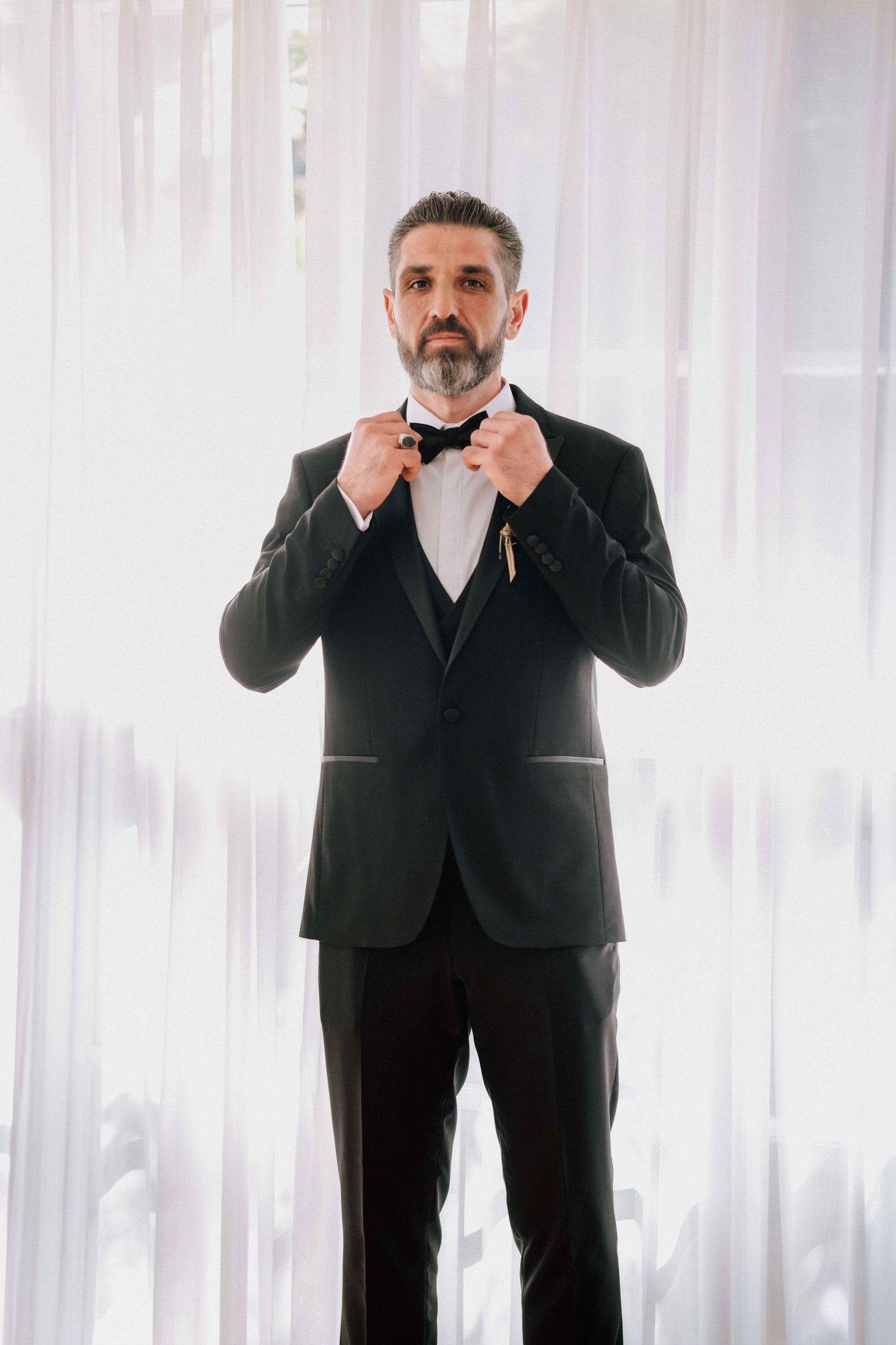 Handsome groom in black tuxedo adjusting bow tie while standing before sheer white curtains.