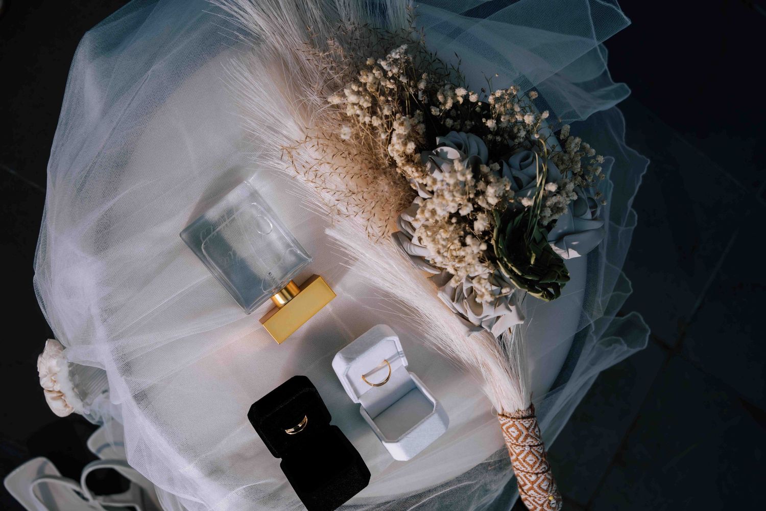Flat lay of wedding accessories including a veil, bouquet, perfume, and ring box on dark surface.