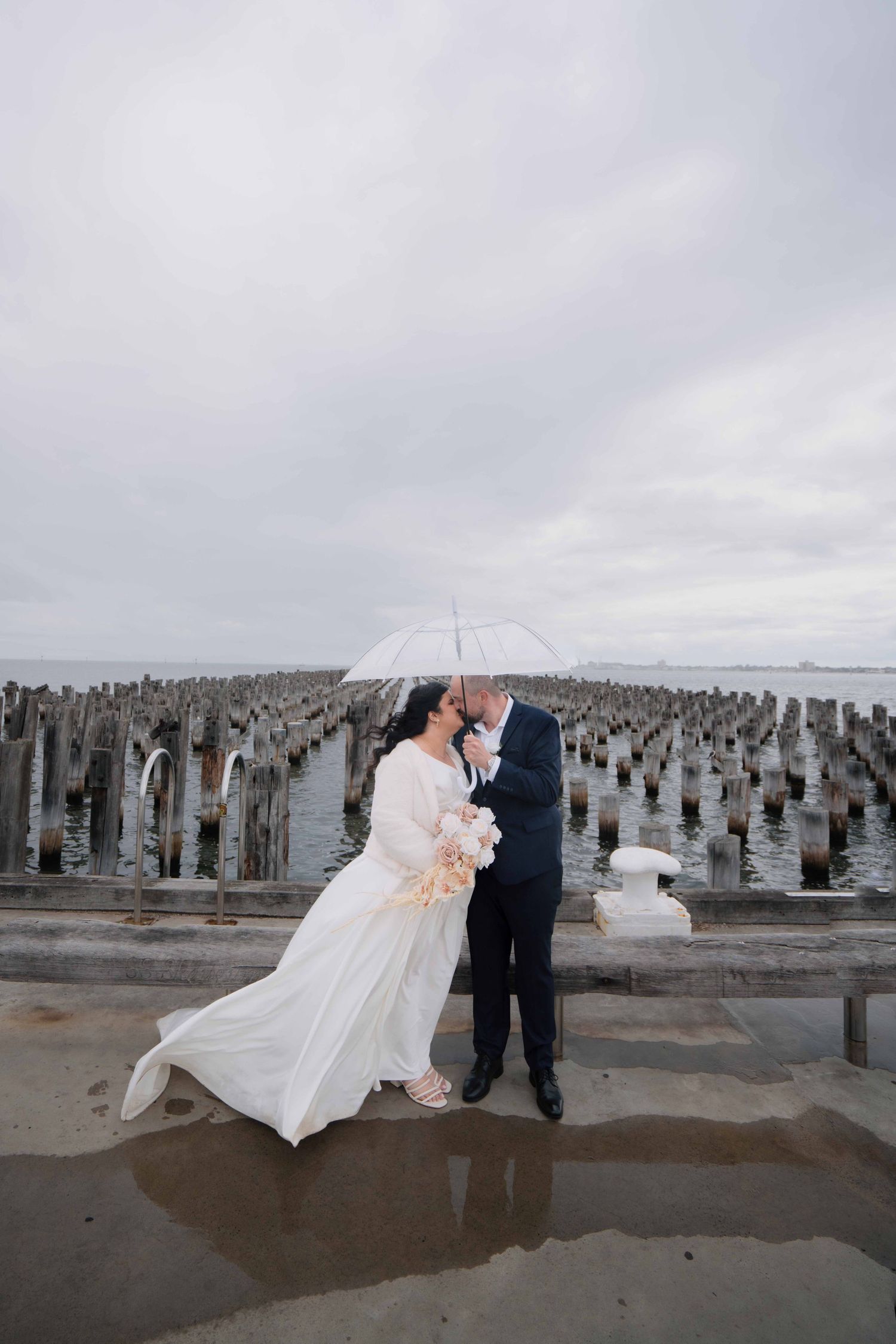 Bride and groom sharing a romantic moment under a clear umbrella at a rainy waterfront pier.