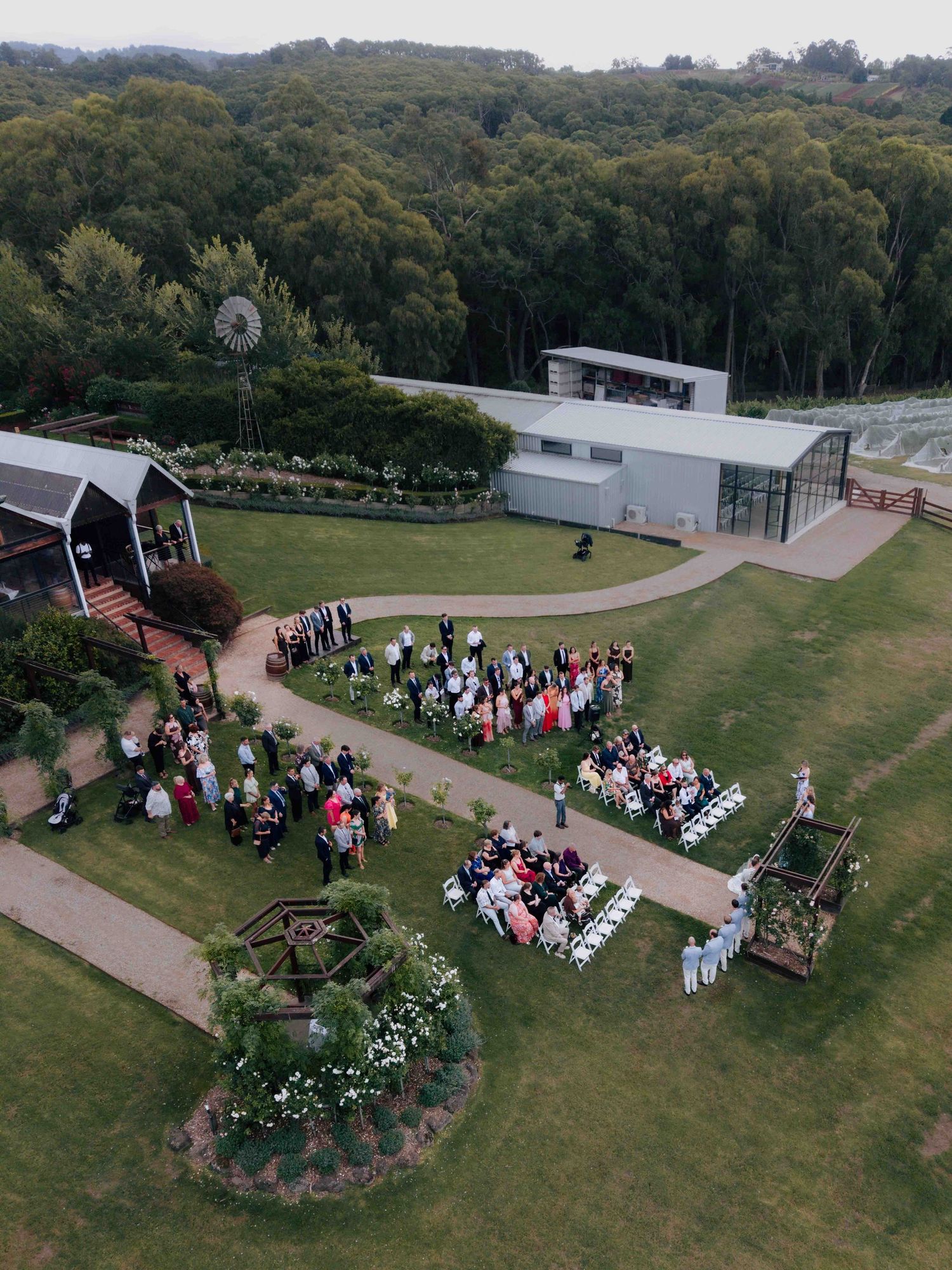Aerial view of outdoor wedding ceremony in progress at a scenic countryside venue with lush greenery.