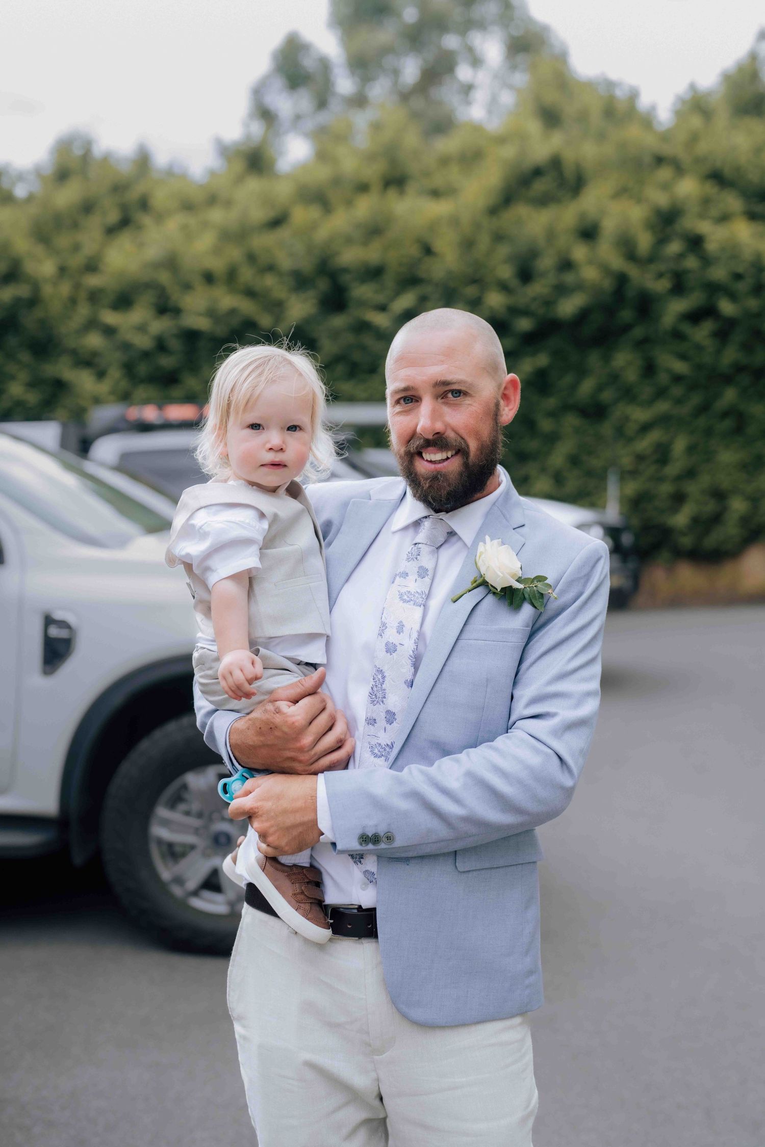 Smiling bald bearded groom in light blue suit holds toddler at outdoor wedding venue.