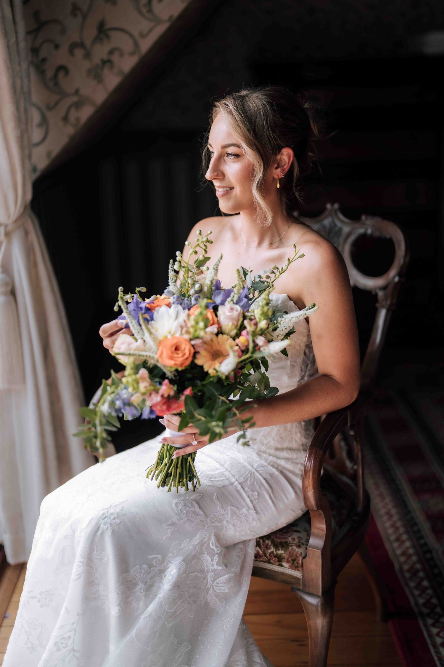 Beautiful bride in white lace wedding dress holding colorful bridal bouquet of orange and purple flowers.