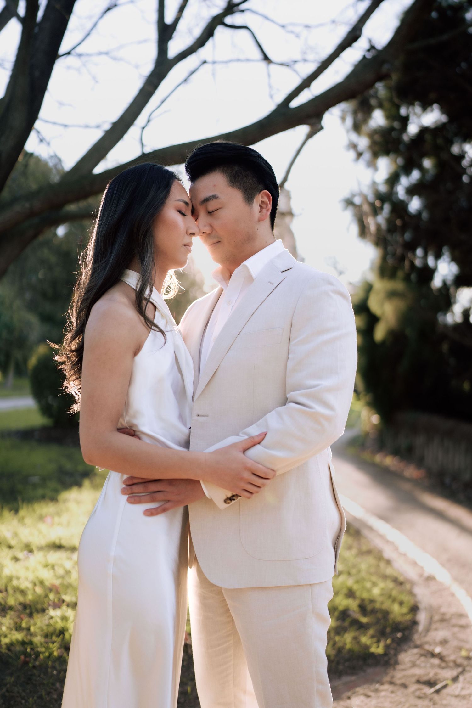 Couple in white outfits sharing a romantic moment in a sunlit garden, with the man in a suit and woman in a sleeveless dress.