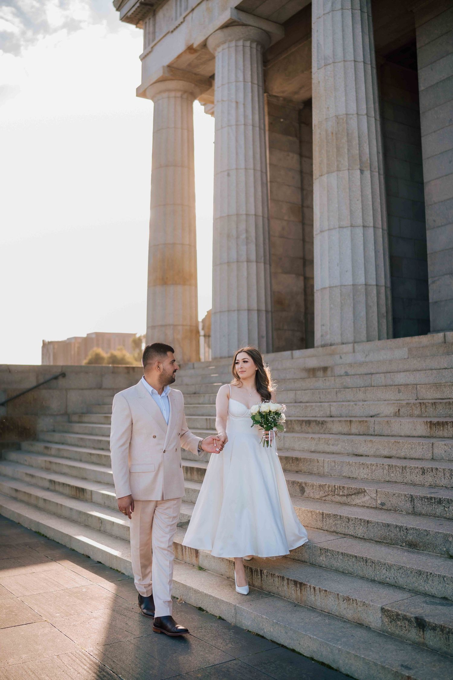 Bride and groom holding hands on sunlit stone steps beside towering classical columns at a grand monument.