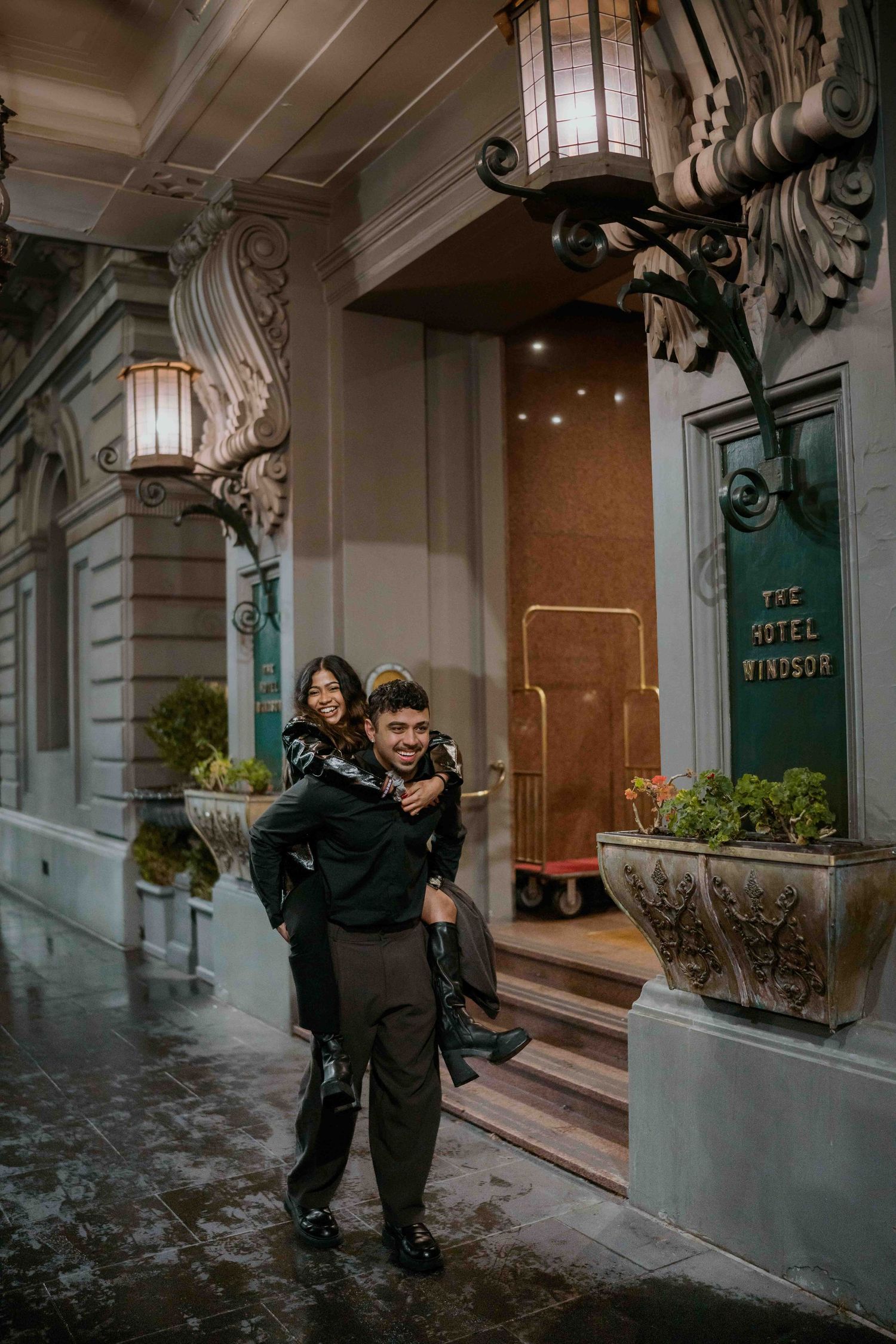Man giving woman a piggyback ride outside The Hotel Windsor entrance on a rainy night.