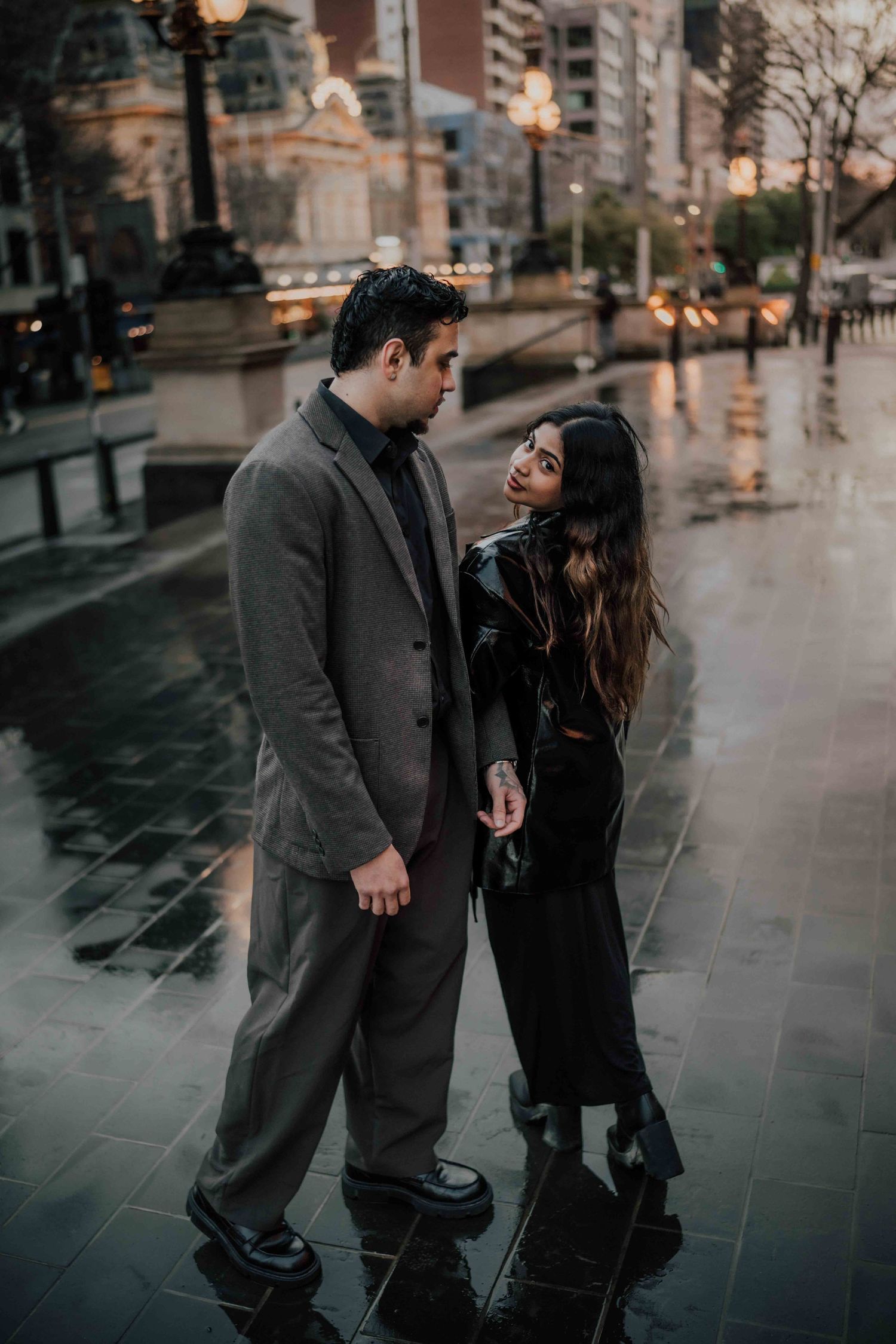 Stylish couple holding hands and sharing a glance on a reflective rainy city street.
