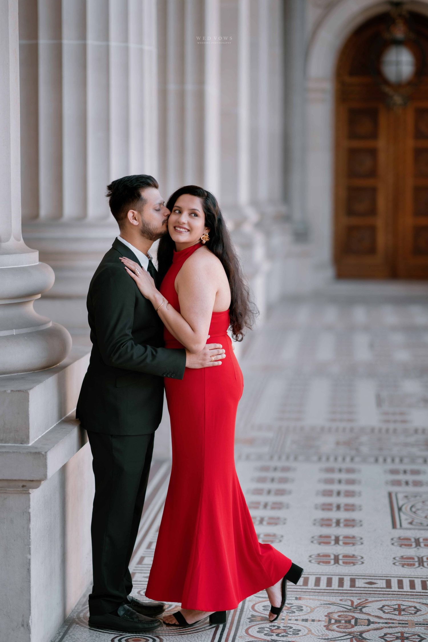 Man in dark suit kissing woman in red halter gown against ornate marble columns in engagement shoot.