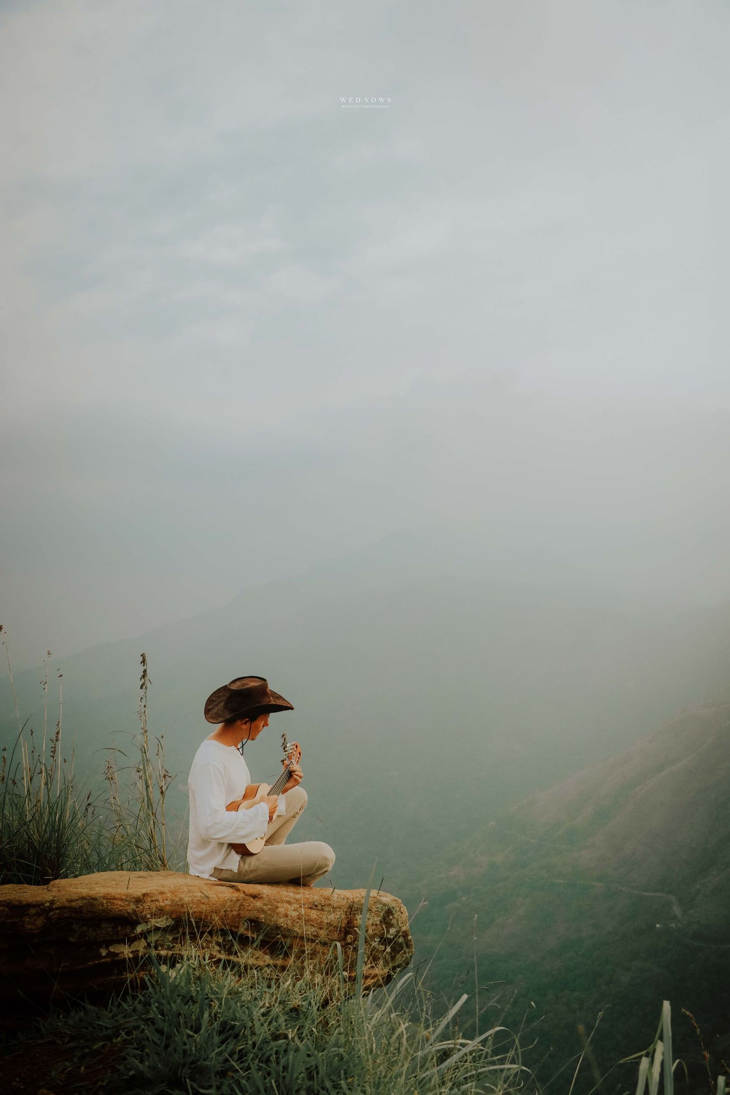 A woman in a wide-brim hat sits cross-legged on a rocky cliff holding wildflowers, surrounded by misty mountain scenery.