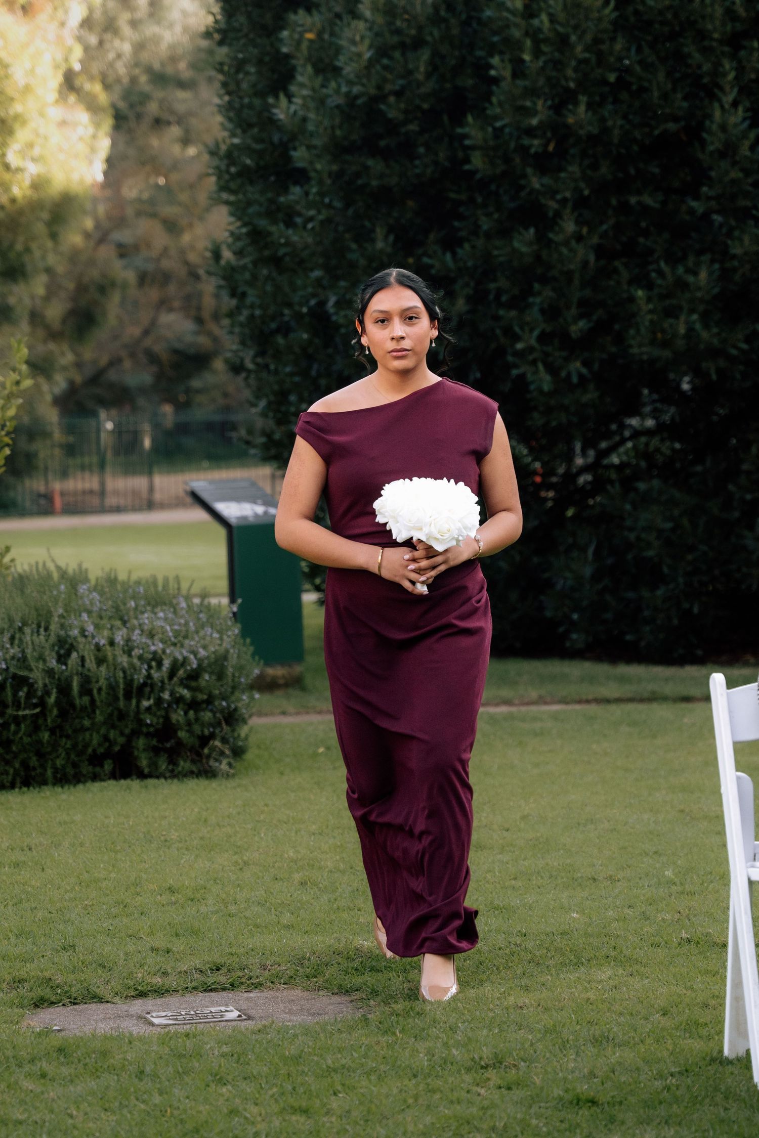 Bridesmaid in burgundy off-shoulder gown walking barefoot down grass aisle holding white floral bouquet.