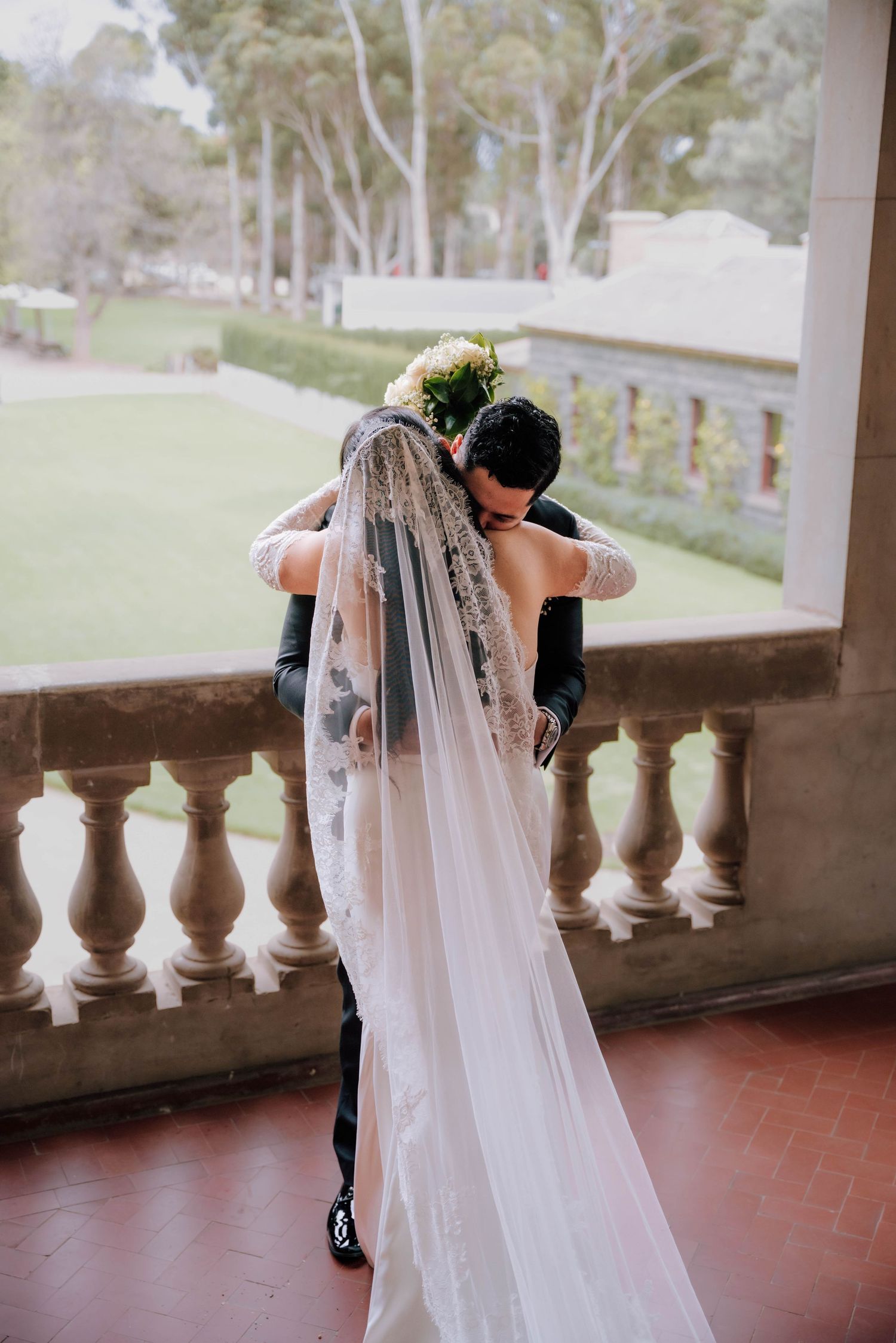 Bride and groom sharing emotional first look embrace on ornate stone balcony with garden views.