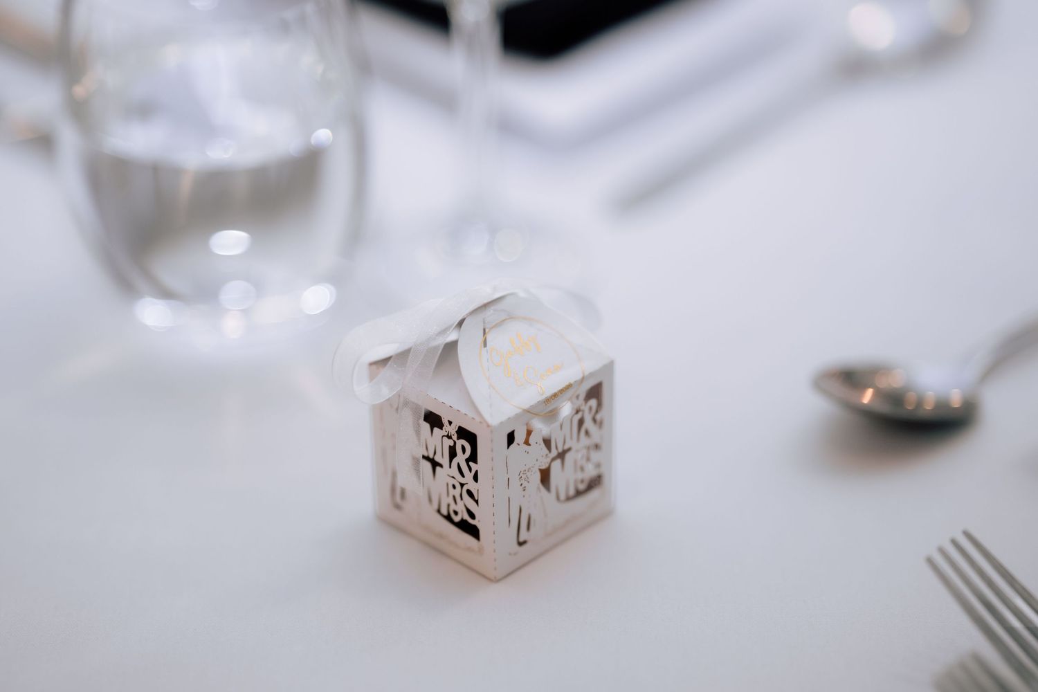 Elegant laser-cut white wedding favor box with ribbon sits on white reception table beside glassware.