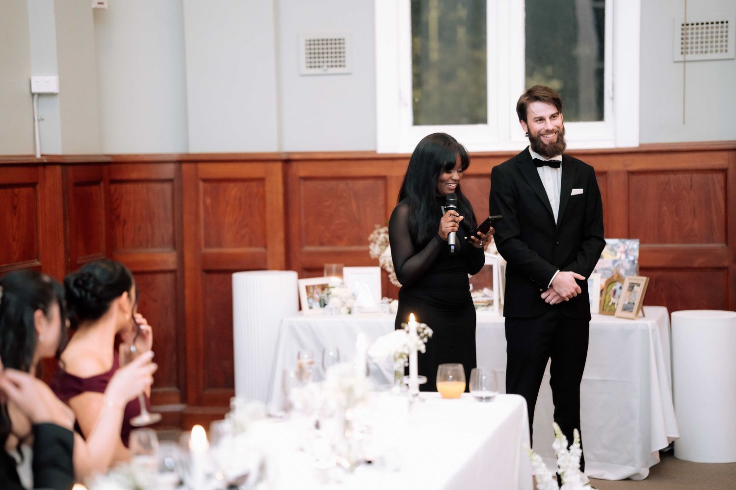 Two wedding speakers deliver a toast at a elegant white-decorated reception hall to seated guests.