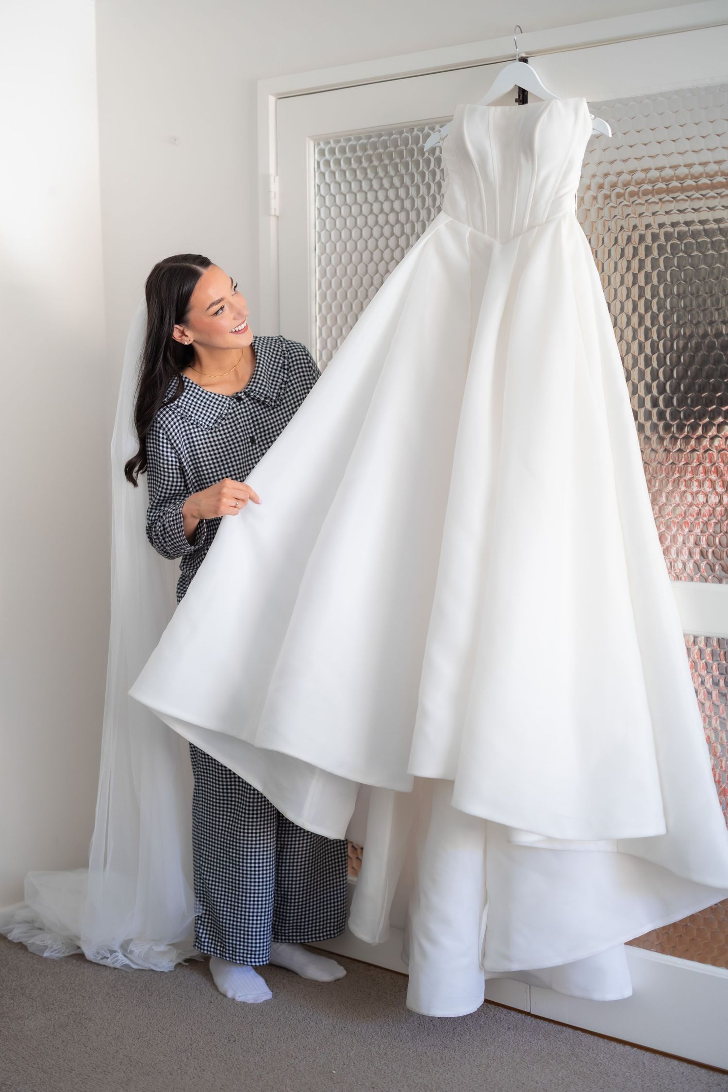 Woman admiring a white ball gown wedding dress hanging on a door in a bright bridal fitting room.