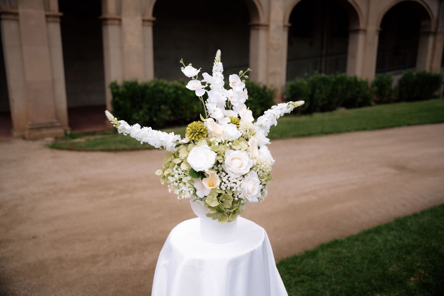 Elegant white floral wedding centerpiece with roses and snapdragons on draped table outdoors.