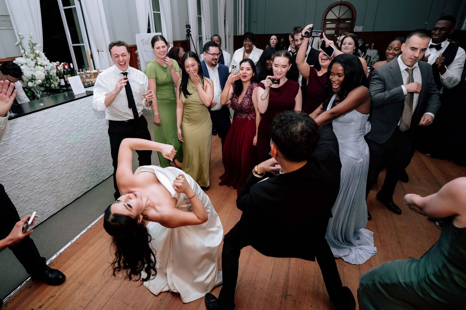 Bride being dipped by groom on crowded wedding reception dance floor surrounded by cheering guests.