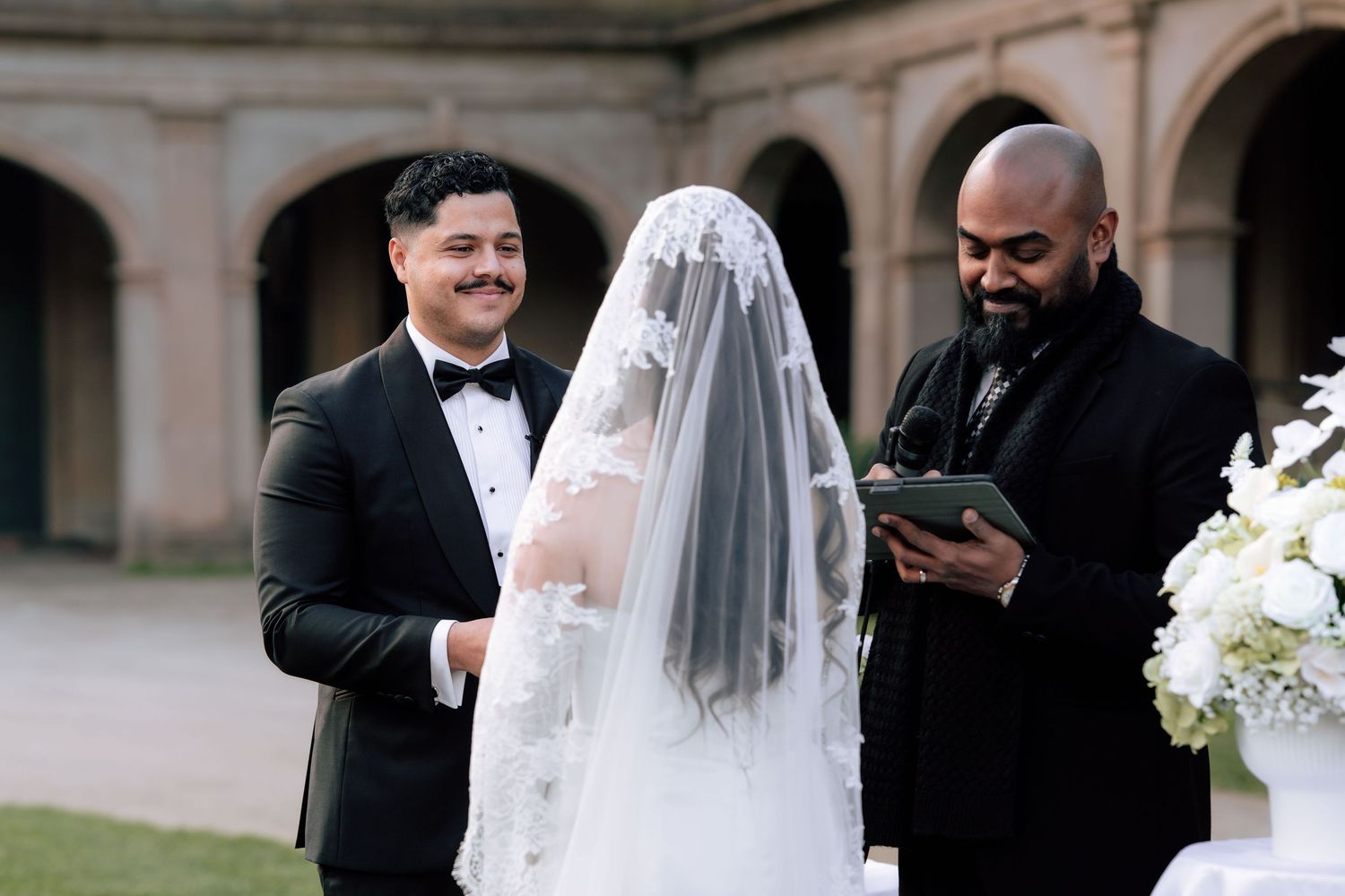 Groom smiling as officiant reads vows while bride in lace veil stands at outdoor wedding ceremony.
