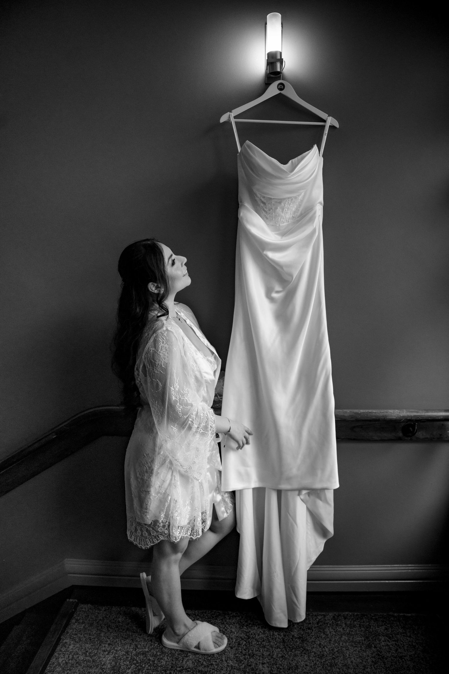 Bride in lace robe admiring her wedding dress hanging on wall in black and white photo.
