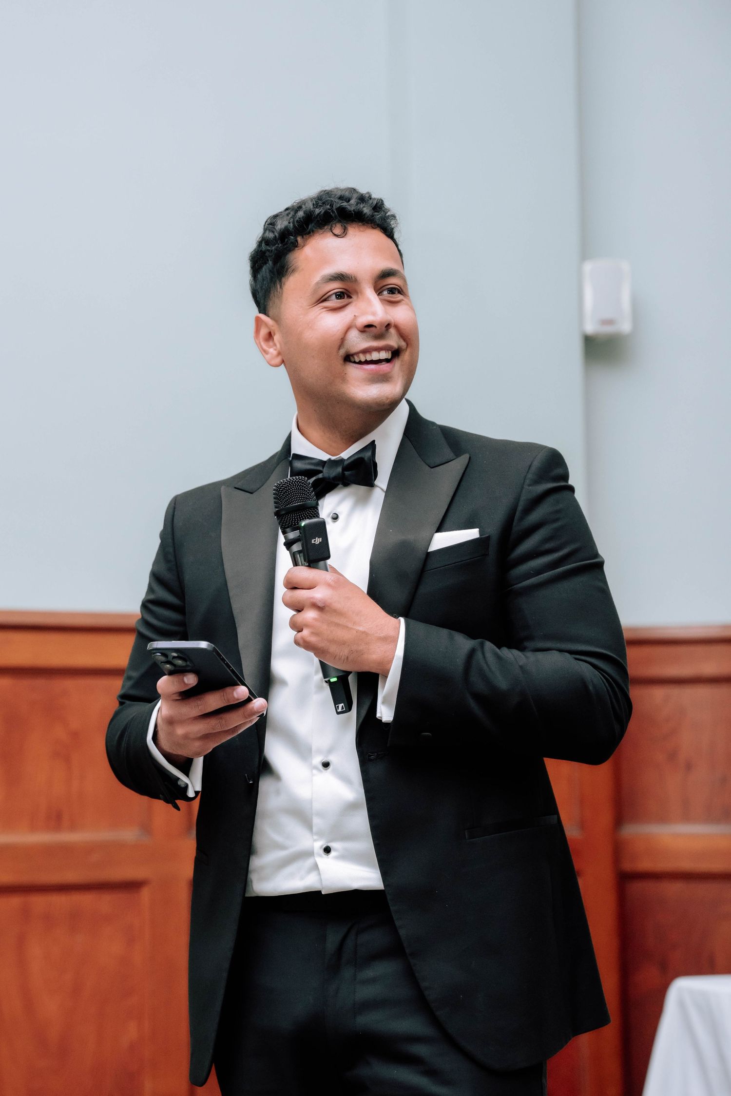 Smiling groomsman in black tuxedo holds microphone and phone while delivering speech at wedding reception.