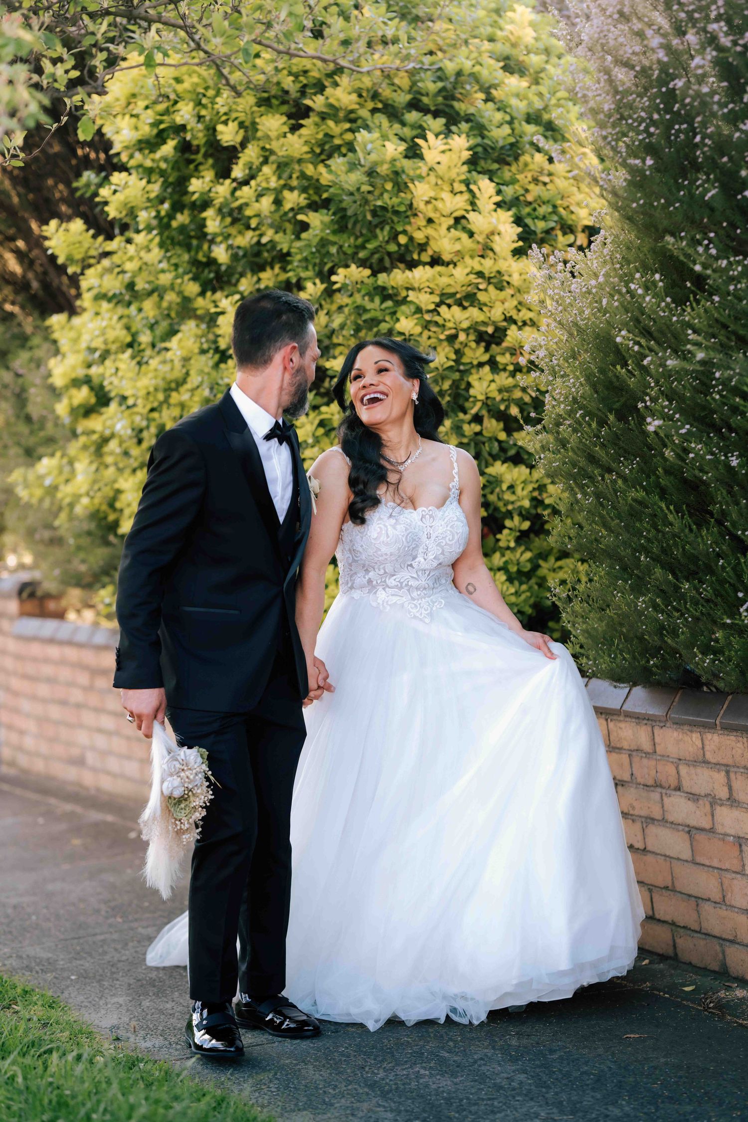 A laughing bride in a white tulle gown holds hands with her groom in a black tuxedo outdoors.