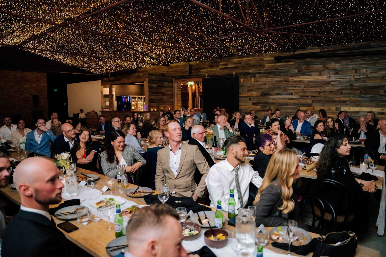 Wedding guests seated at long tables watching a presentation under warm fairy light ceiling.