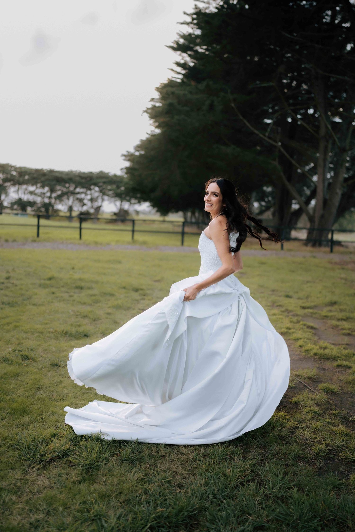 Bride twirling in flowing white wedding gown on green grass with trees in background.