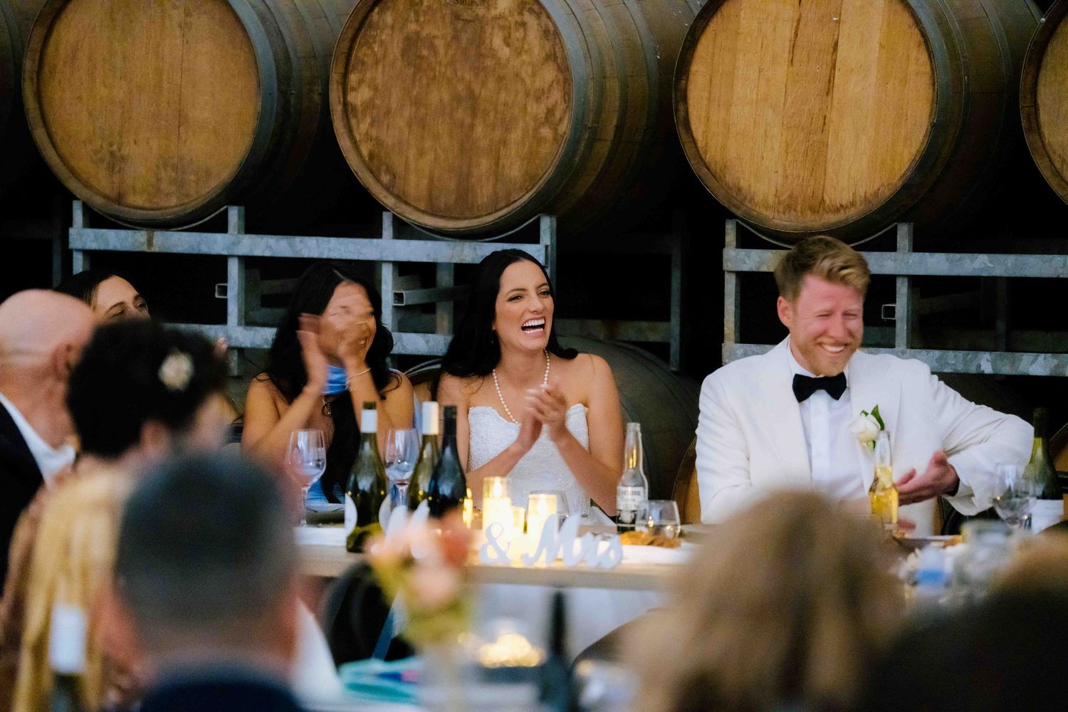 Bride in white gown laughs with groom in tuxedo at head table in front of wine barrels.