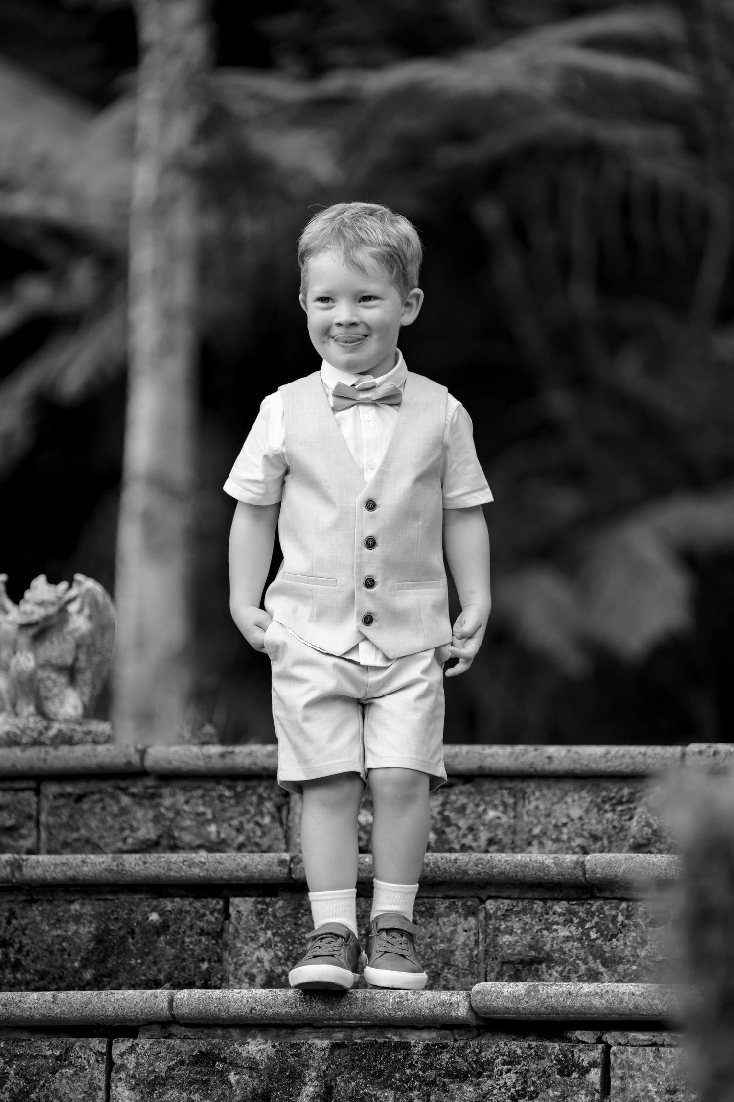 Adorable young boy in cream vest and shorts with bow tie smiling while standing on stone wall.